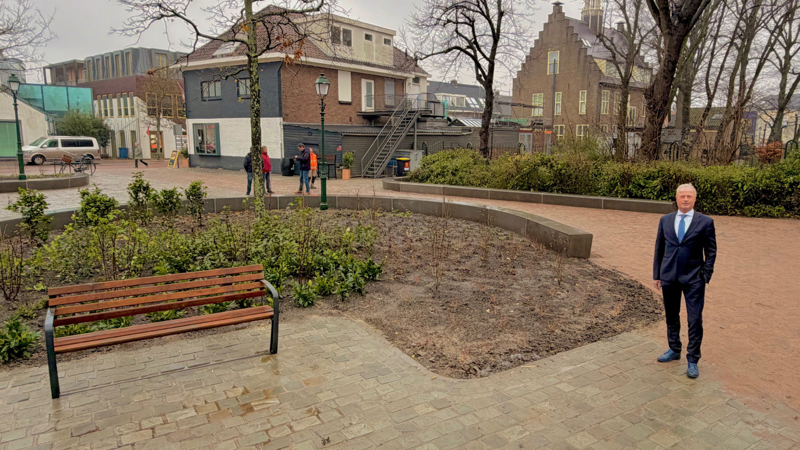 Een man in een pak staat op een plein met bankjes en struiken, omgeven door gebouwen en bomen in een regenachtige dag.