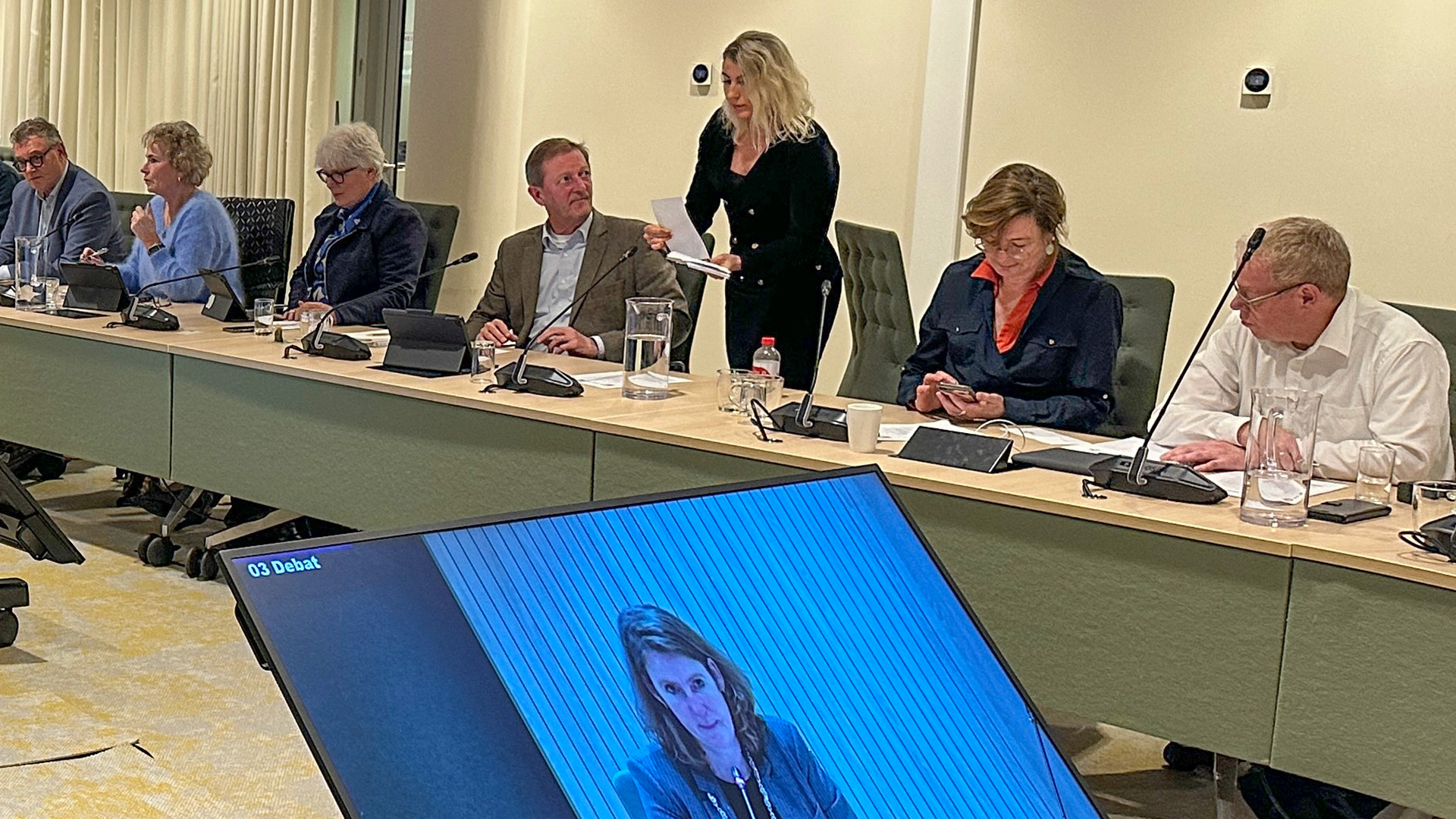 Mensen zitten rond een conferentietafel tijdens een vergadering, met een scherm dat een spreker toont en "03 Debat" aangeeft.