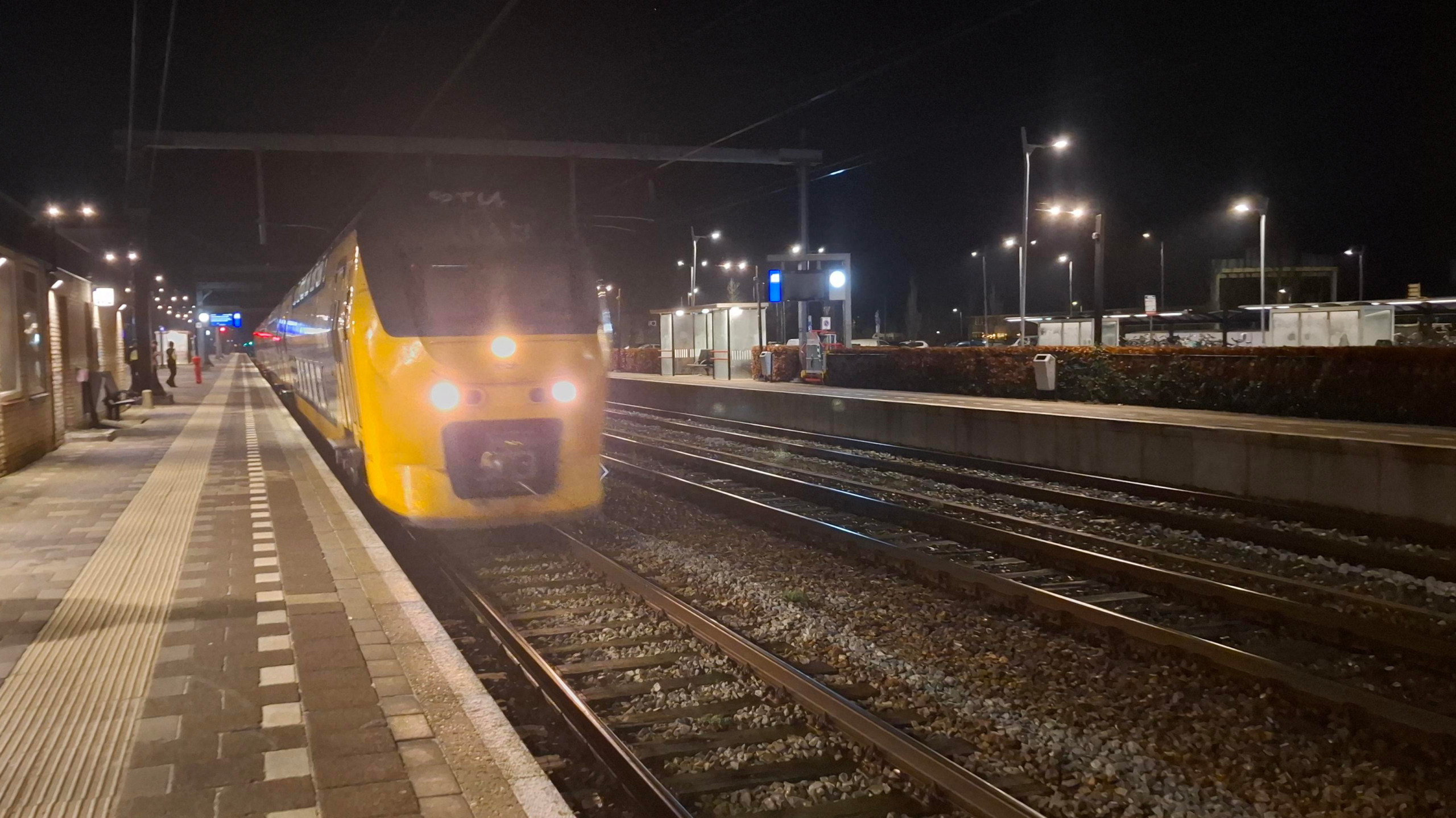 Gele dubbeldeks trein arriveert 's nachts op een verlicht perron met enkele wachtende reizigers.