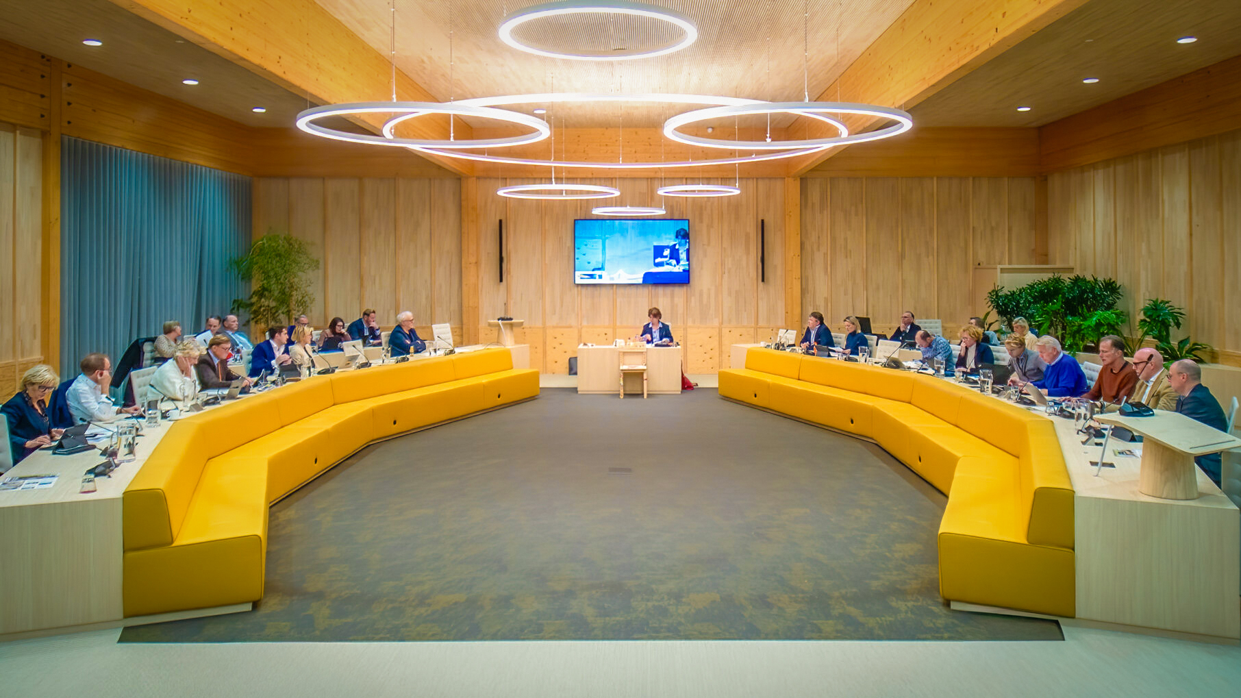Mensen vergaderen in een moderne zaal met houten interieur en gele banken, onder grote ronde lampen.