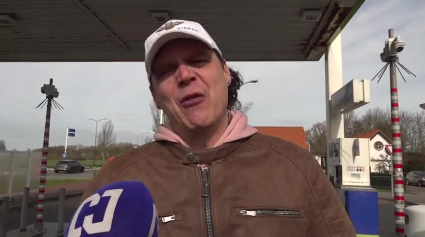 Man met witte pet en bruine jas spreekt in een microfoon bij een benzinestation, opname overdag.