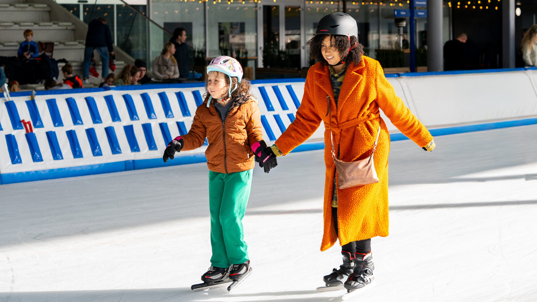 Een vrouw in een oranje jas helpt een kind met schaatsen op een ijsbaan.