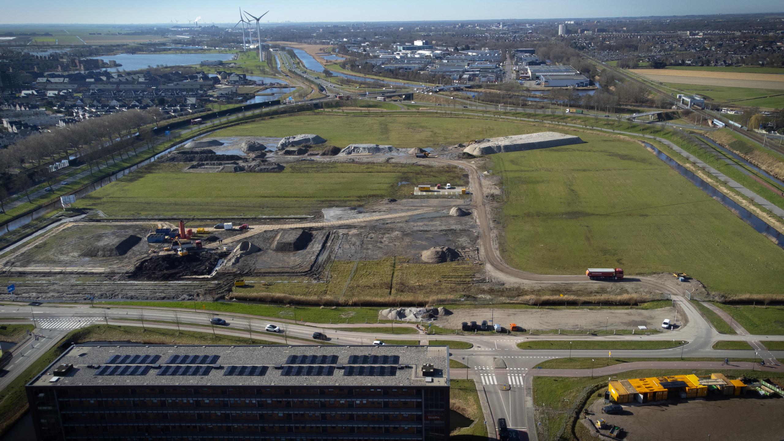 Luchtfoto van een bouwterrein met machines en voertuigen, omgeven door groene velden en wegen, met windmolens en gebouwen op de achtergrond.