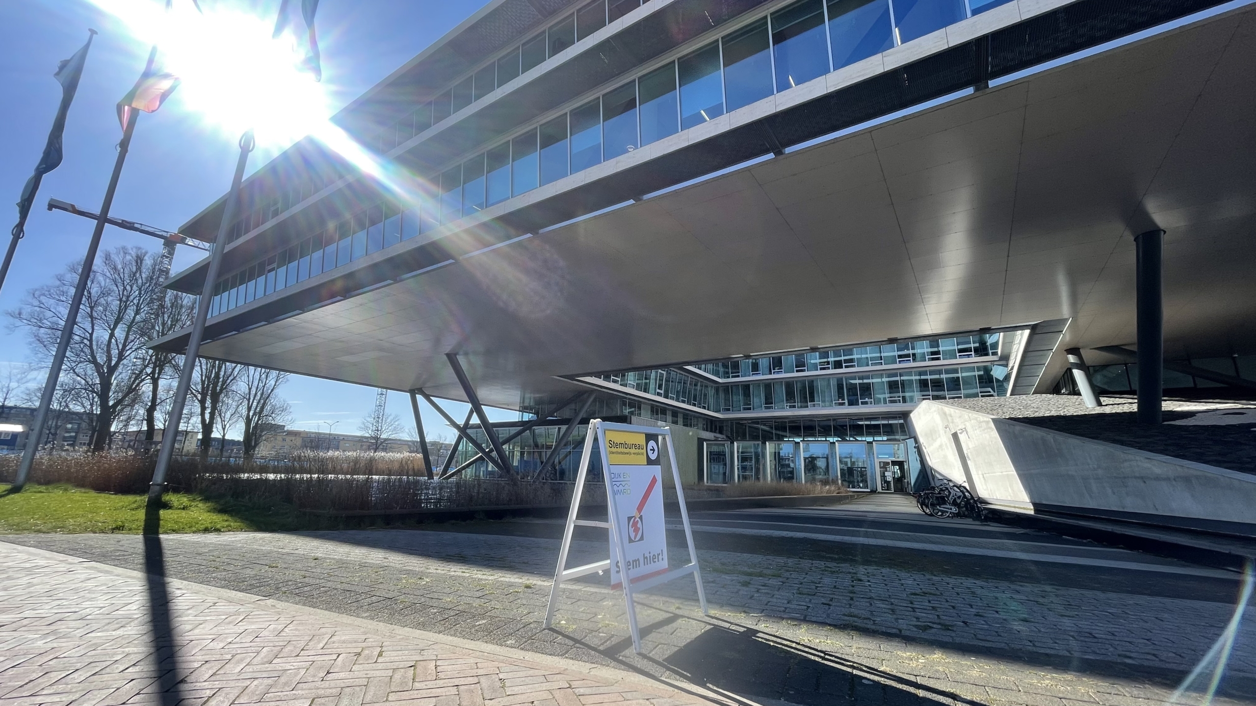 Een modern gebouw met grote ramen en een bord dat een stembureau aanwijst onder een zonnige hemel.