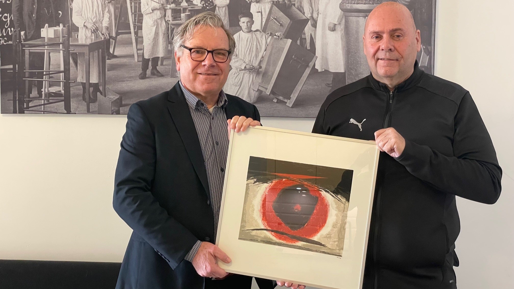 Twee mannen houden een kunstwerk met een rood en zwart abstract ontwerp vast, voor een zwart-wit fotoachtergrond.