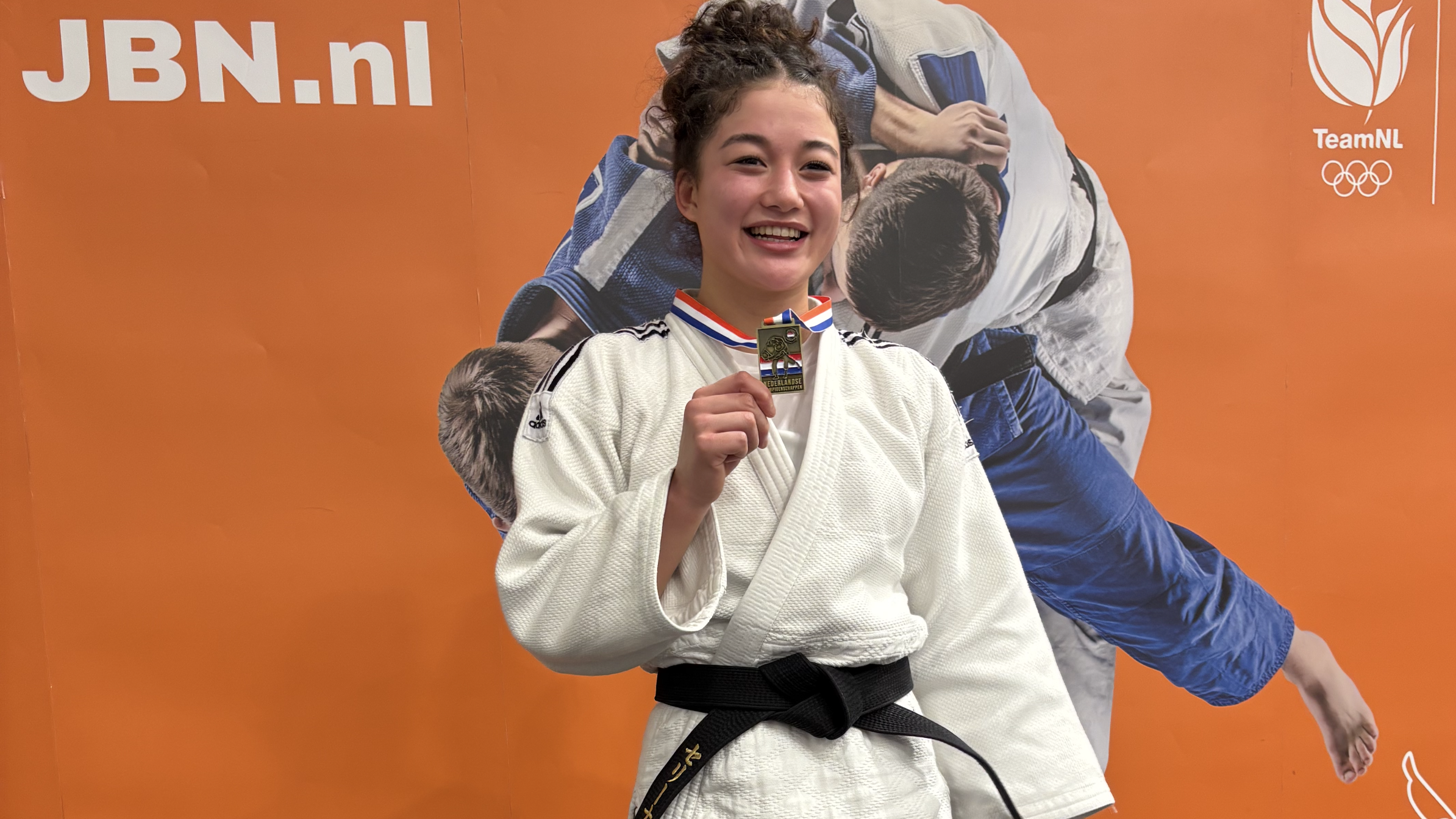 Judoka in judopak houdt glimlachend een medaille vast voor een oranje achtergrond met logo's.