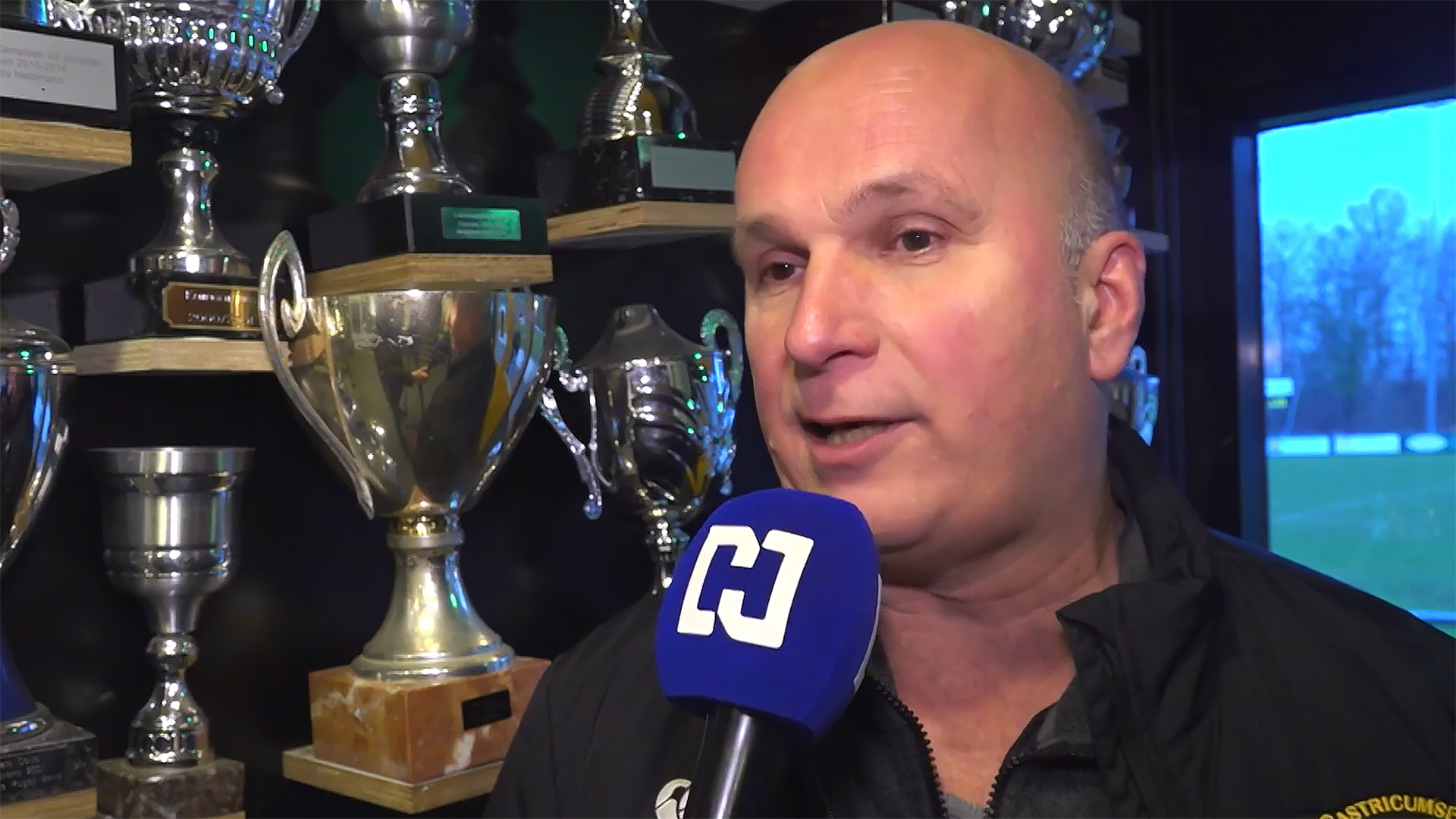 Man geeft een interview met trofeeën op de achtergrond en een blauwe microfoon met logo in beeld.