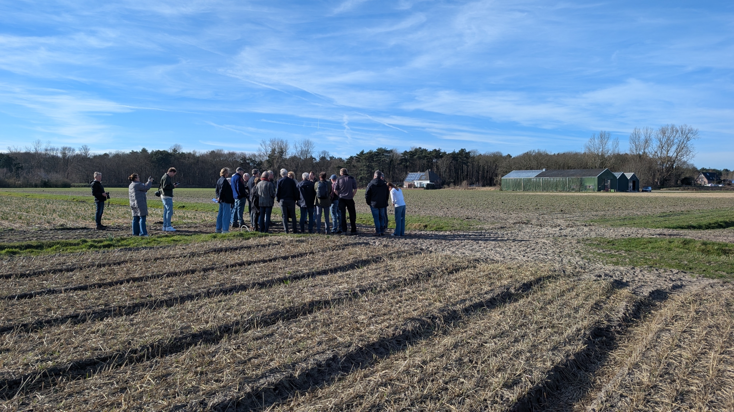 Een groep mensen staat op een akker bij een kas en een boerderij, onder een blauwe lucht.