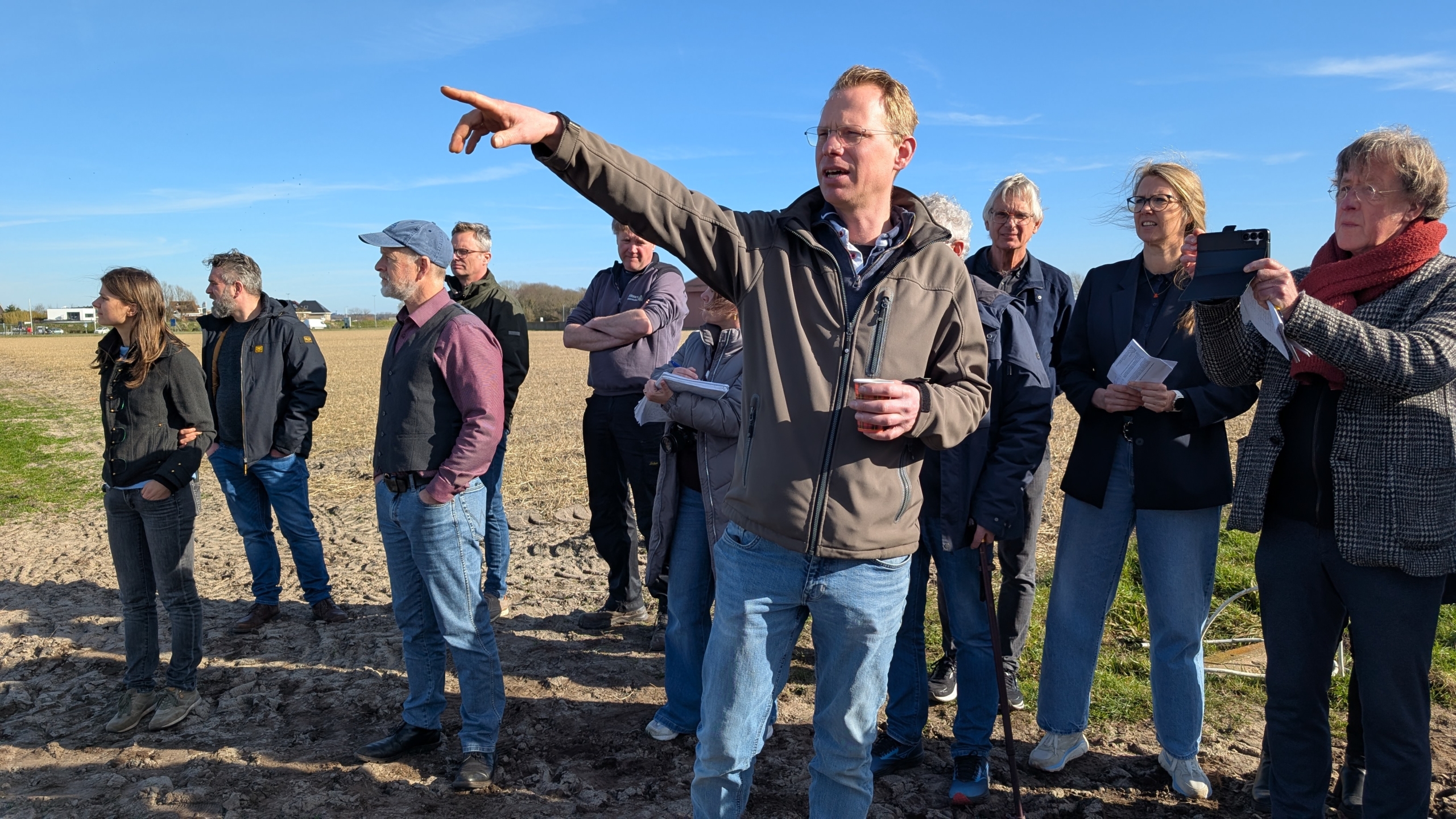 Een man wijst iets aan in de verte terwijl hij met een groep mensen op een open veld staat.