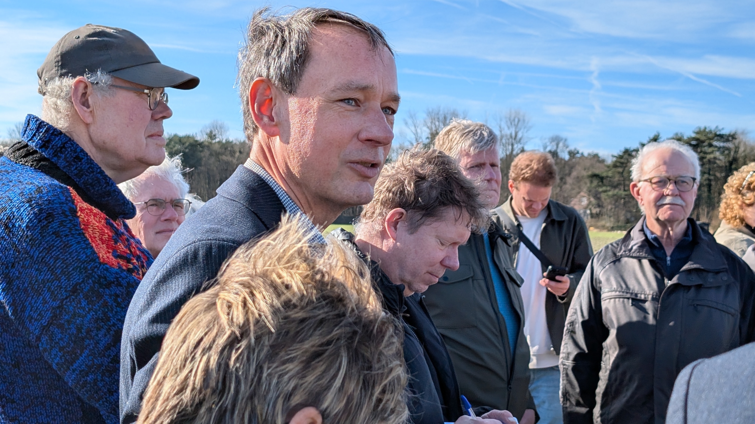 Een groep mensen staat buiten bijeen onder een heldere hemel, kijkend in verschillende richtingen.
