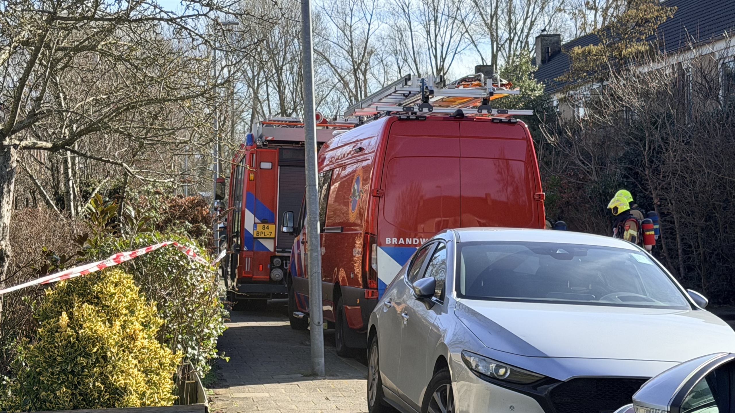 Brandweerwagens en een brandweerman in een woonwijk, met afzetlint en geparkeerde auto's op de voorgrond.