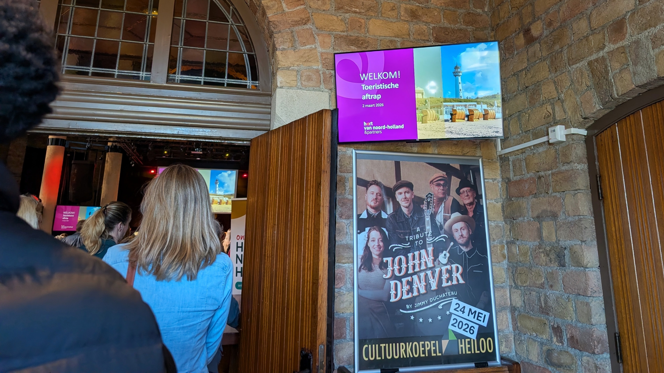 Mensen bij de ingang van een evenement met een scherm dat "Welkom! Toeristische aftrap" toont en een poster voor een John Denver tribute.