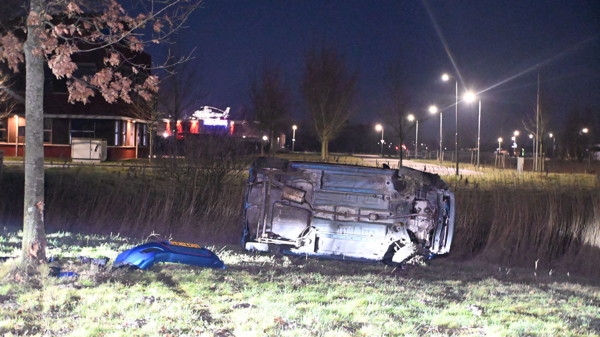 Auto ligt op zijn zij in het gras langs een weg, omgeven door straatlantaarns en een gebouw op de achtergrond, 's nachts.
