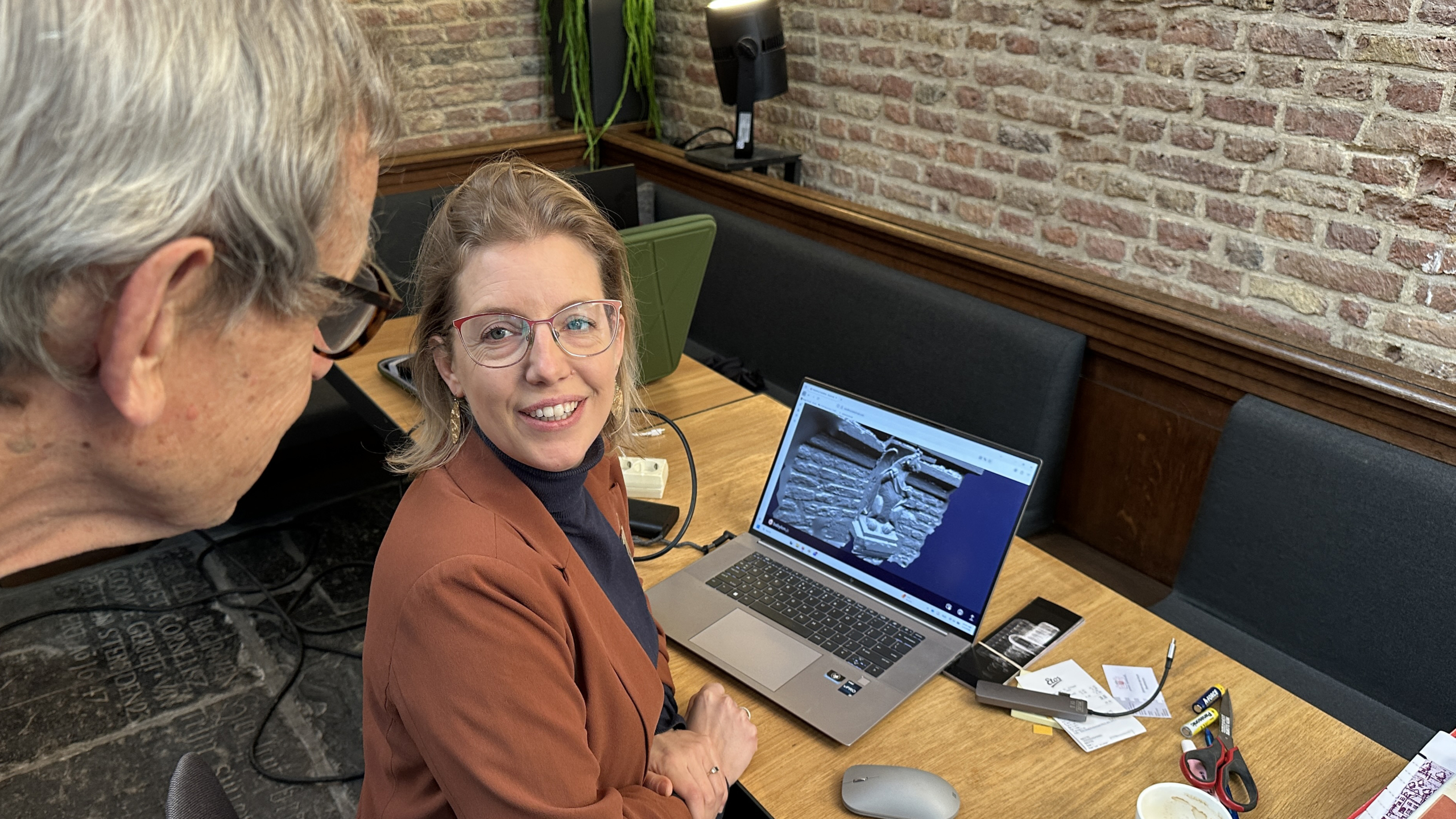 Een vrouw zit aan een tafel met een open laptop waarop een afbeelding is te zien, terwijl een man met grijs haar naar het scherm kijkt.