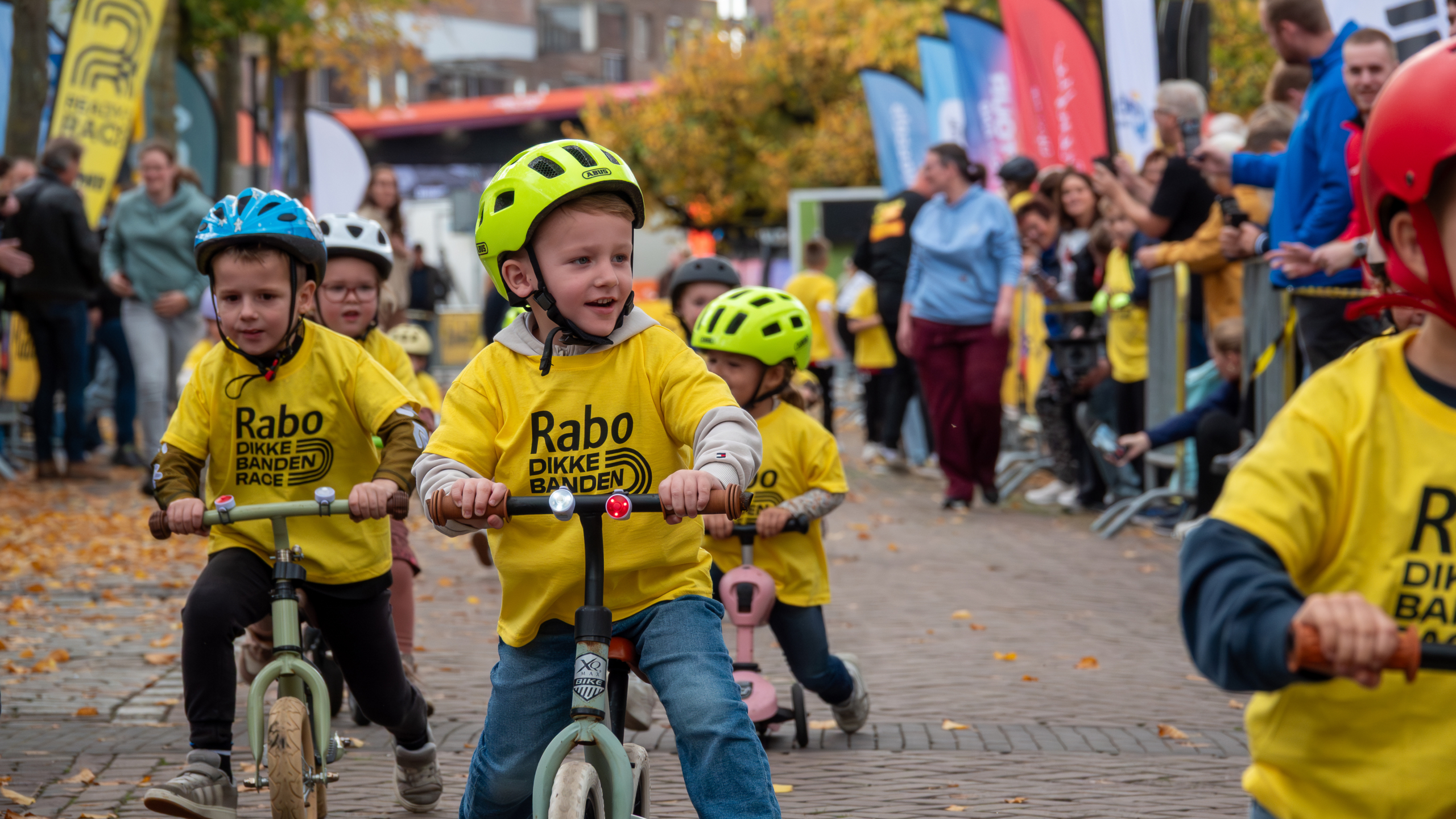 Kinderen met gele helmen en shirts nemen deel aan de Rabo Dikke Banden Race, omringd door toeschouwers.