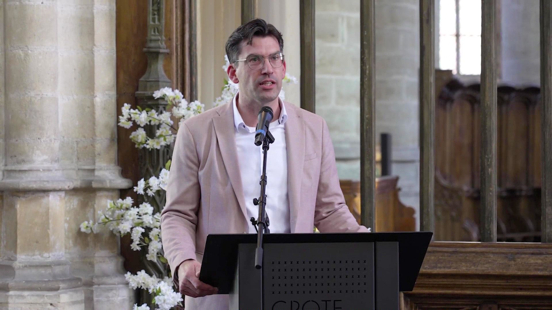 Man in een lichtgekleurde blazer spreekt in een microfoon voor een lessenaar in een sfeervolle kerkelijke omgeving.