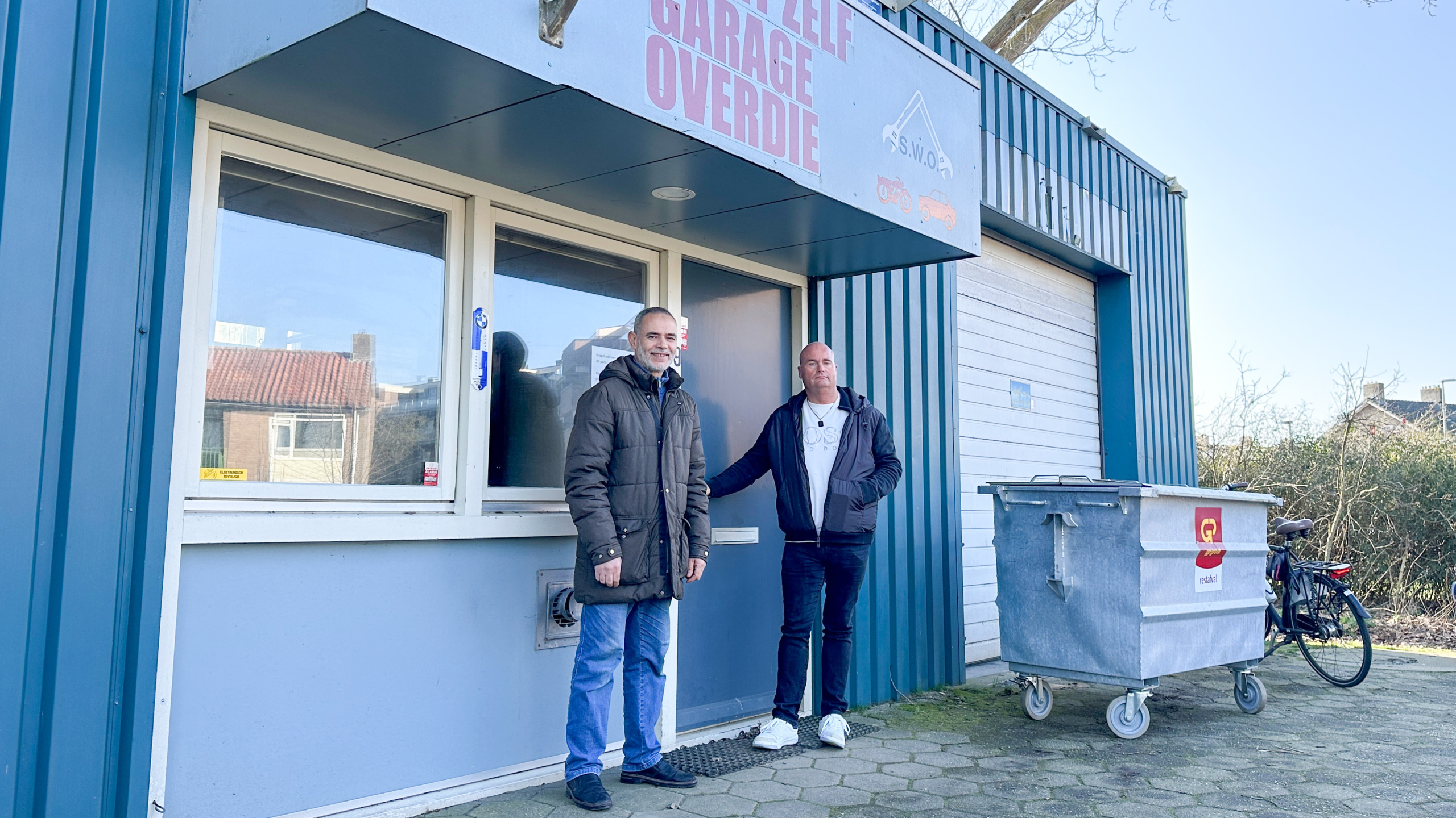 Twee mannen staan voor een garage met het bord "Zelfgarage Overdie". Er is een afvalcontainer en een fiets naast hen.
