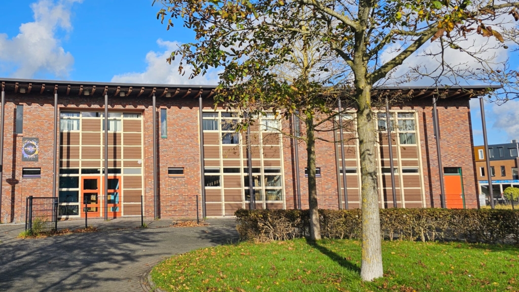 Bakstenen gebouw met grote ramen, omgeven door gras en een boom. De lucht is helder en blauw met enkele wolken.