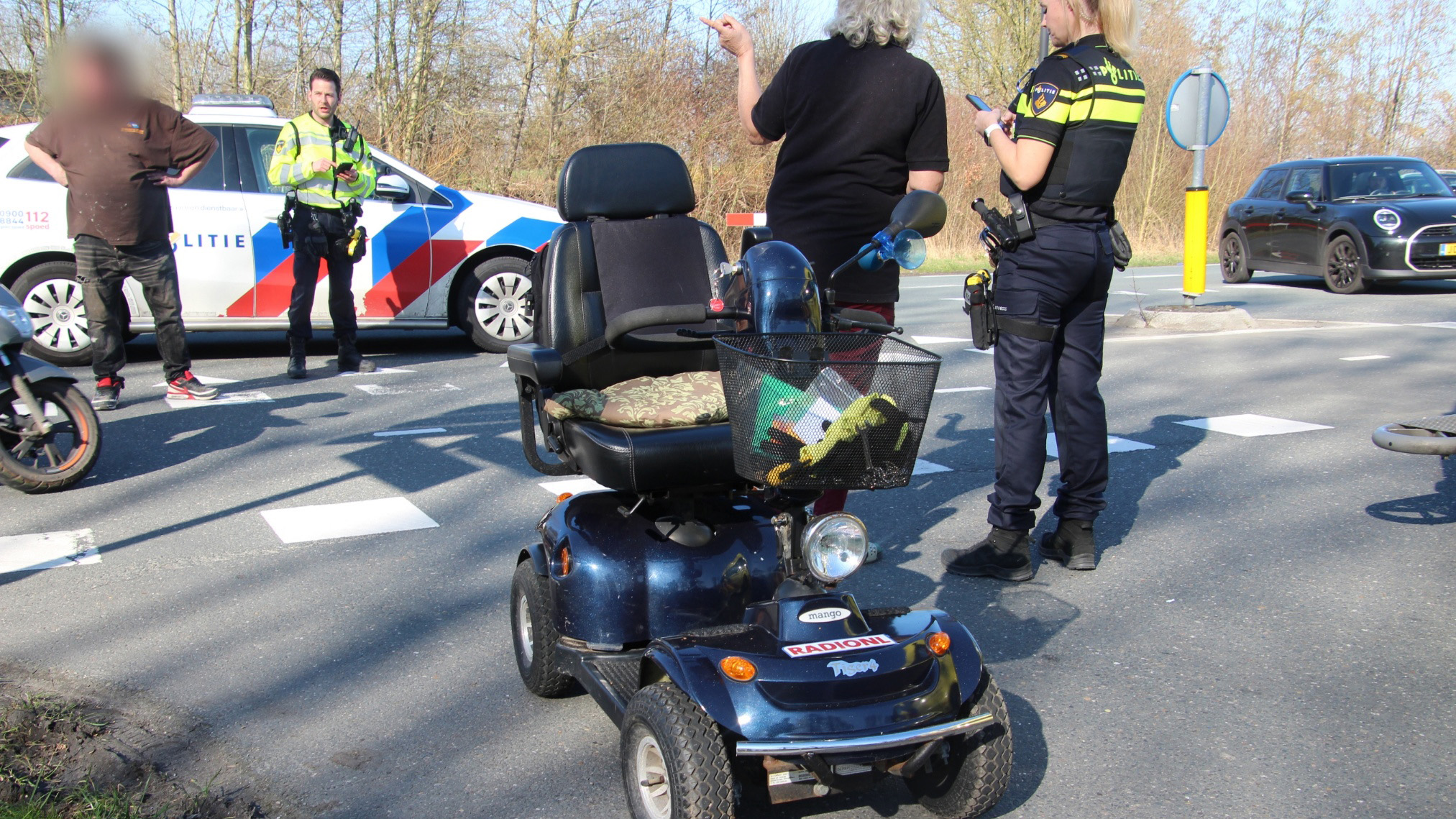 Scootmobiel op een kruispunt met politieagenten en een politiewagen op de achtergrond.