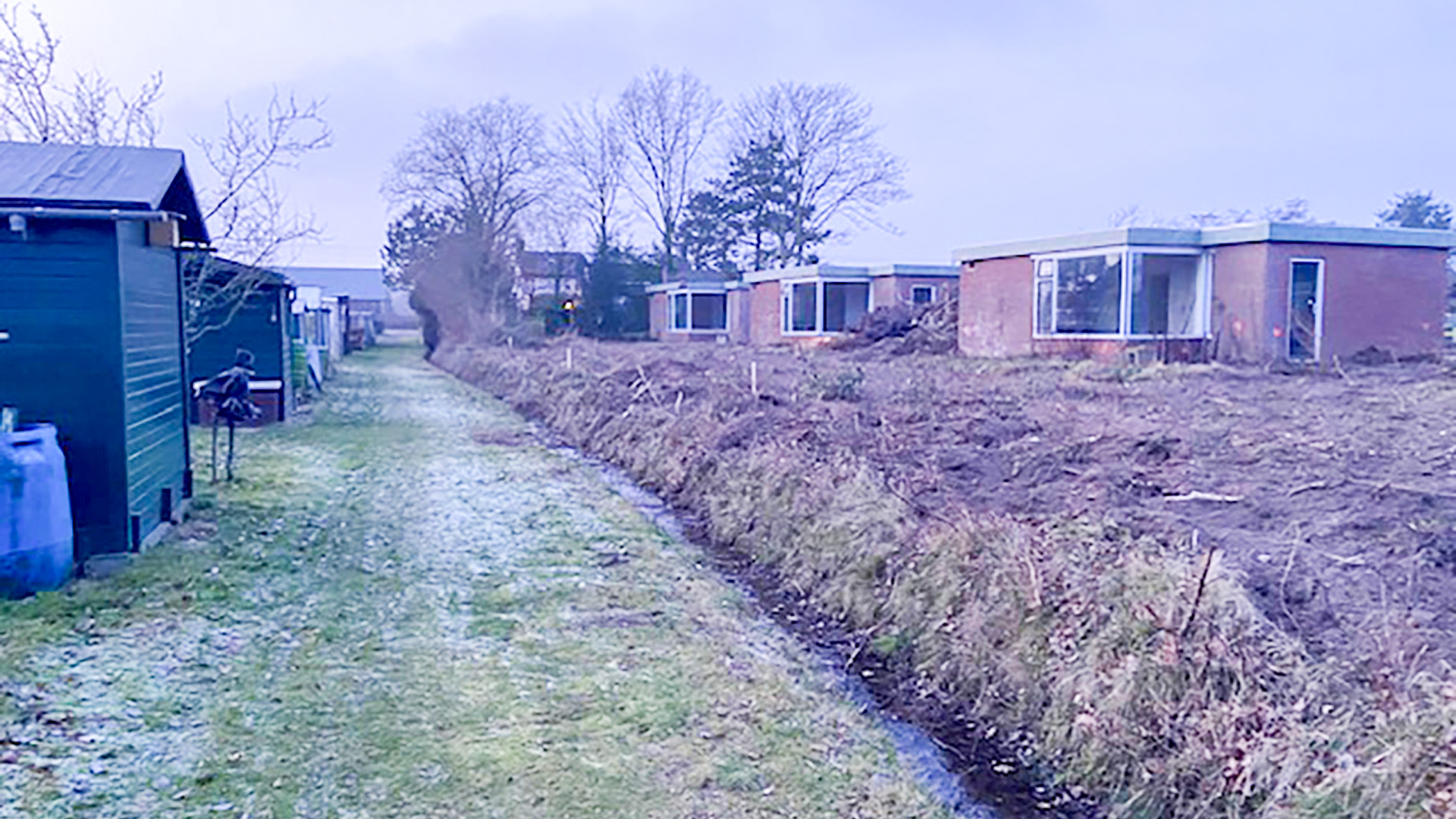 Rij van laagbouw huizen langs een onverhard pad met gras en kale bomen in een winterse setting.