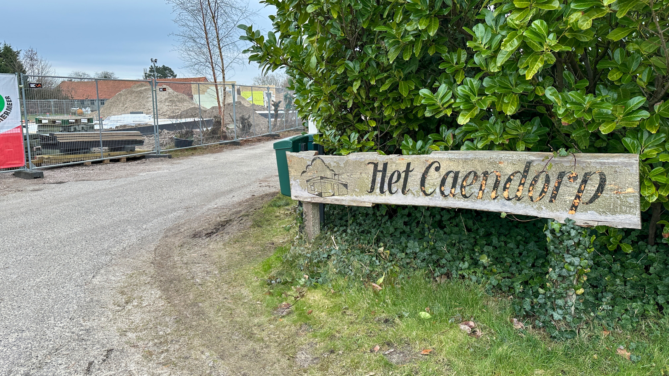 Houten bord met de tekst "Het Caendorp" voor groene struiken, naast een bouwterrein met hekken en stapels materialen.
