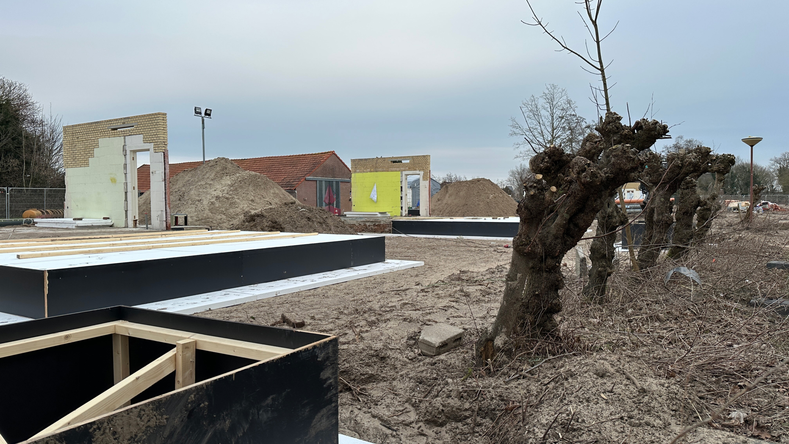 Bouwplaats met gedeeltelijk afgebroken muren en funderingen in voorbereiding, met op de achtergrond stapels aarde en geknotte bomen.