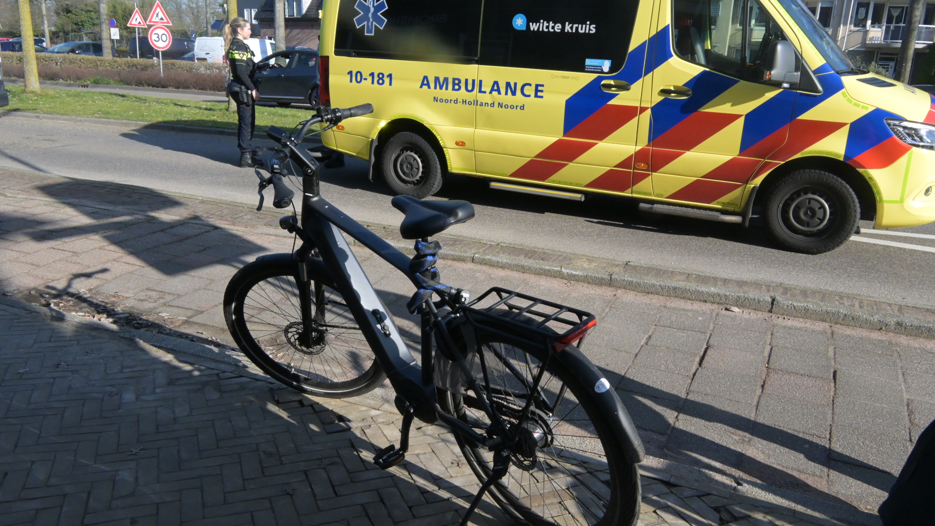 Een geparkeerde fiets op de stoep voor een ambulance en verkeersbord met een politieagent op de achtergrond.