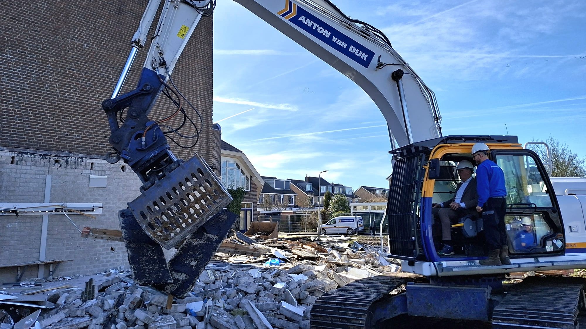 Graafmachine met grijper sloopt een muur; twee mannen in de cabine dragen veiligheidshelmen.