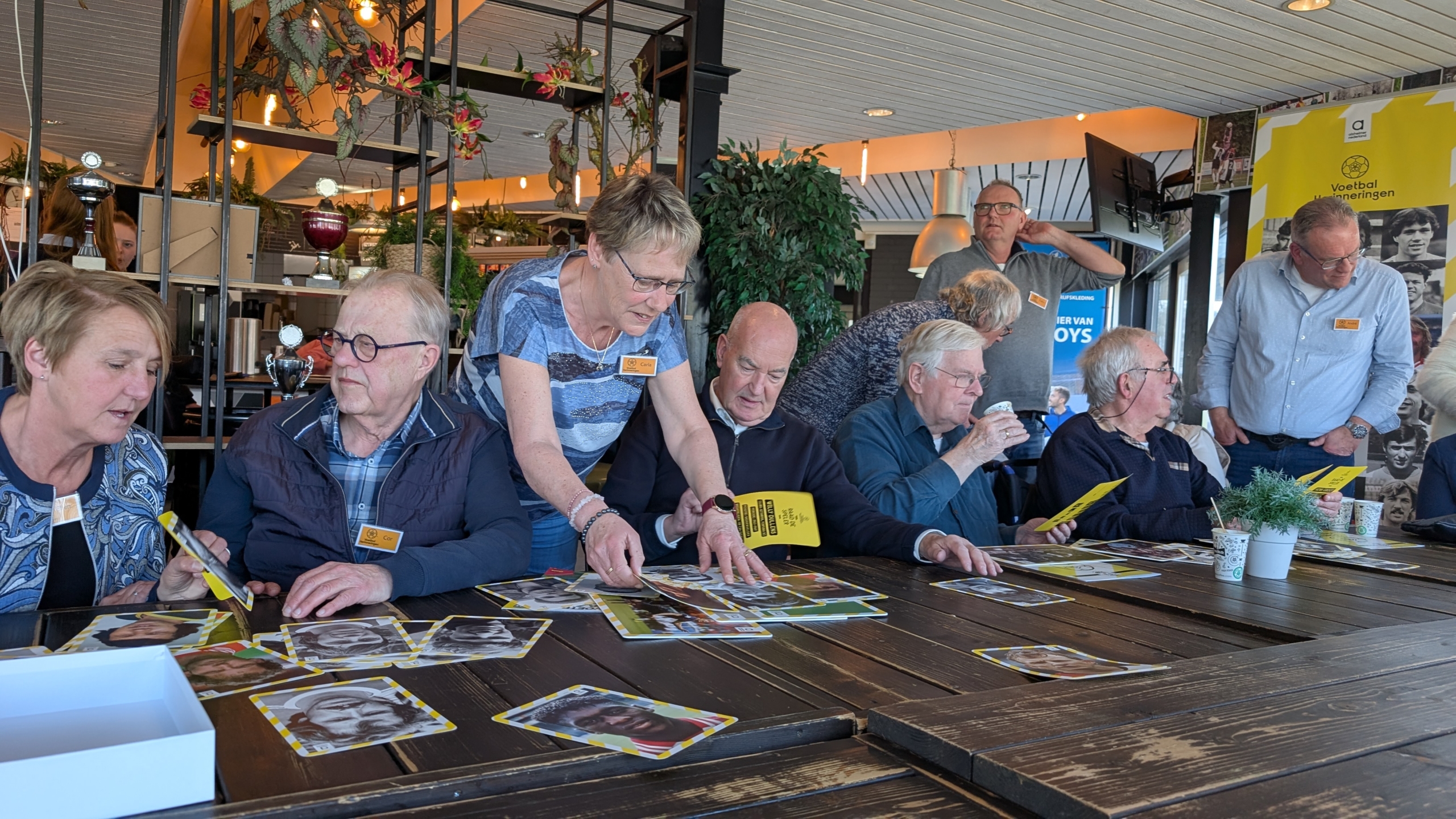 Groep mensen in een café-achtige setting, bezig met het bekijken van foto's en documenten, met planten en bekers op tafel.