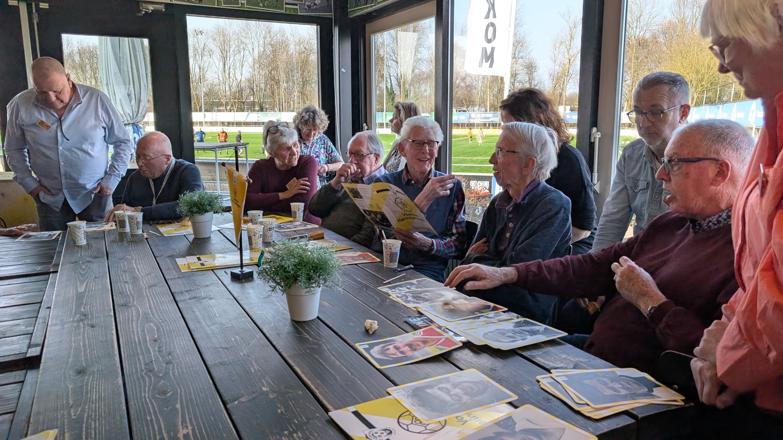 Een groep oudere mensen zit aan een tafel in een sportclubhuis, pratend en foto's bekijkend, terwijl buiten een veld zichtbaar is.