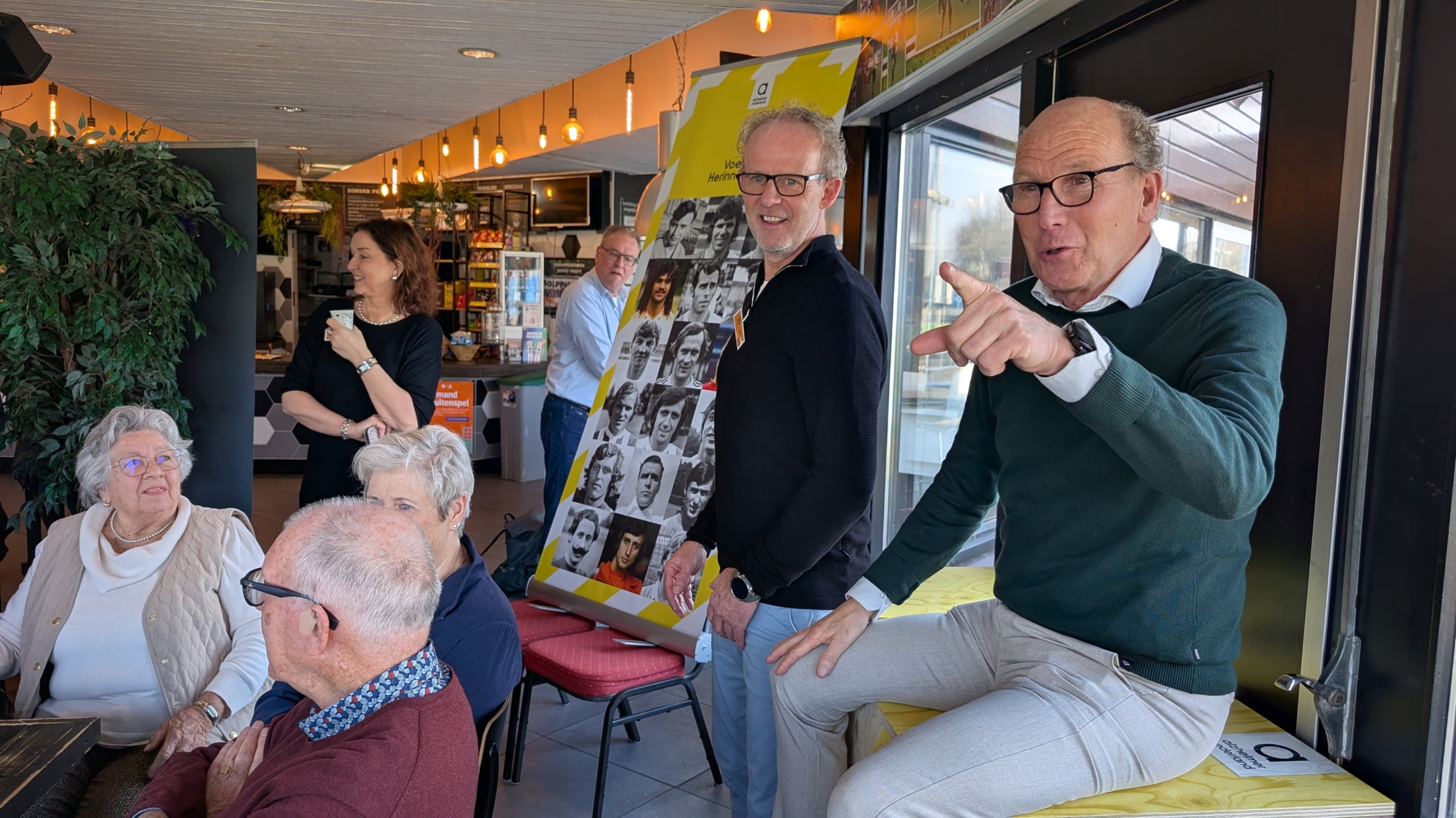 Een groep mensen in een café, met een man die wijst naar de camera en een poster met zwart-wit foto's op de achtergrond.