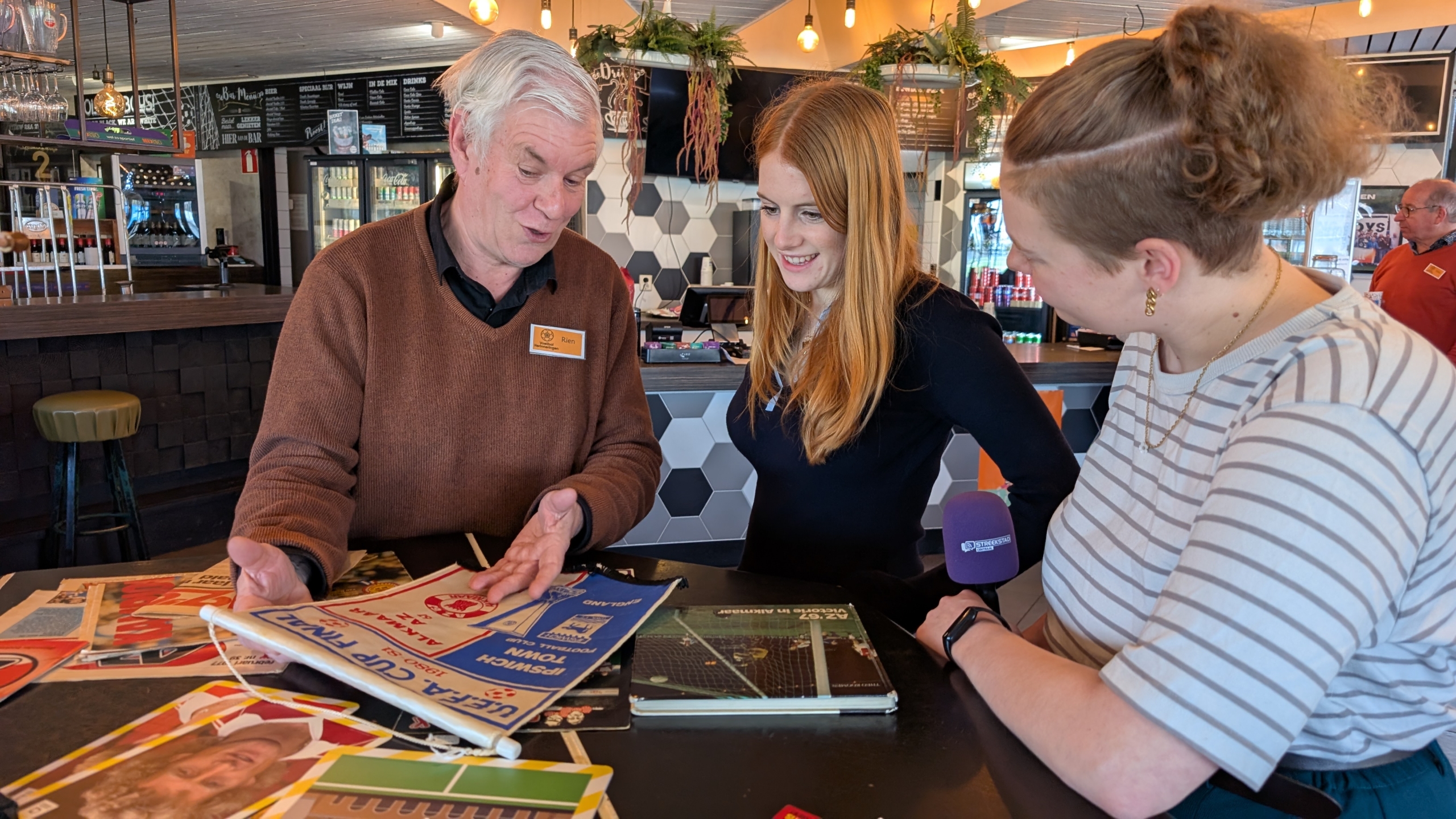 Drie mensen bekijken samen oude voetbalprogramma's in een caféachtige omgeving.