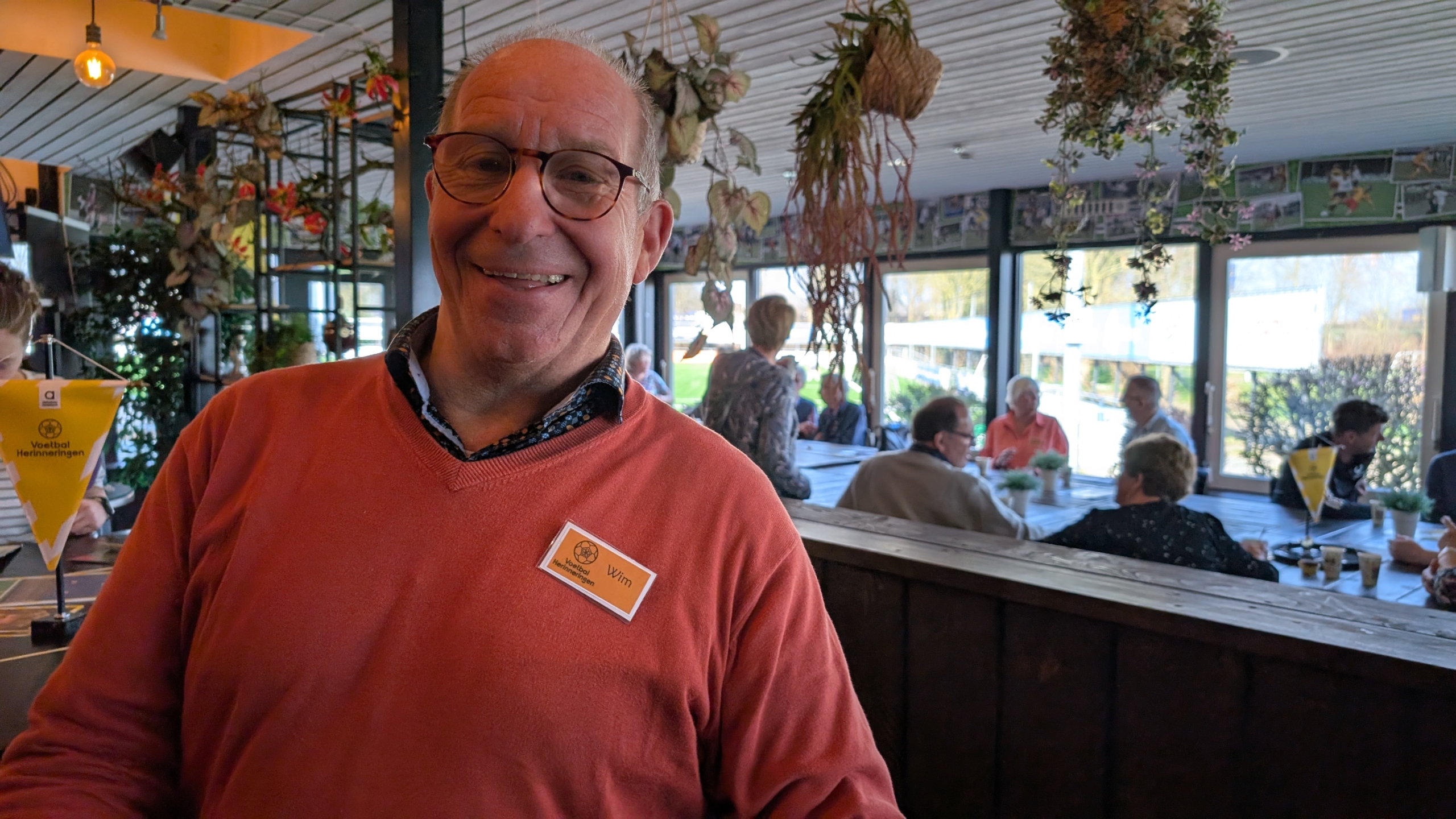 Glimlachende man met een naamkaartje, in een ruimte met planten waar mensen aan tafels zitten.