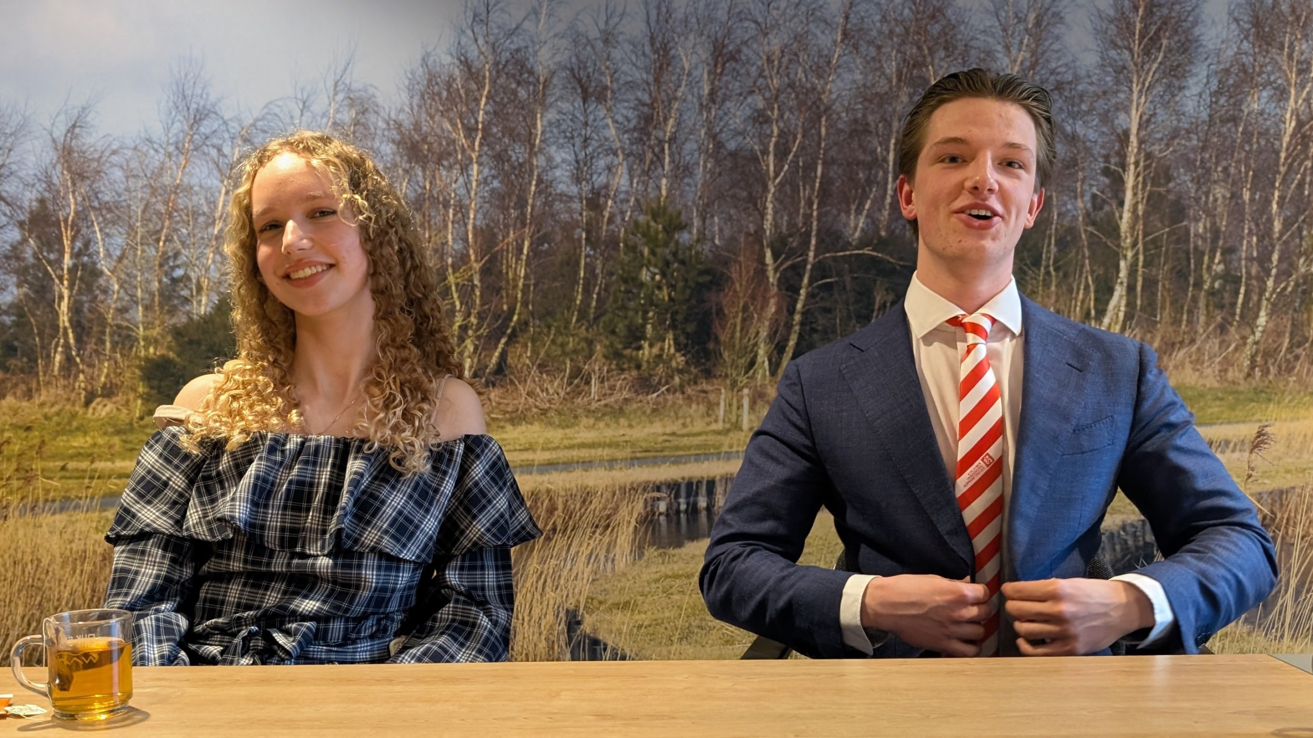 Twee jonge mensen zitten aan een tafel met een natuurachtergrond; de vrouw glimlacht en de man draagt een blauw pak met gestreepte stropdas.