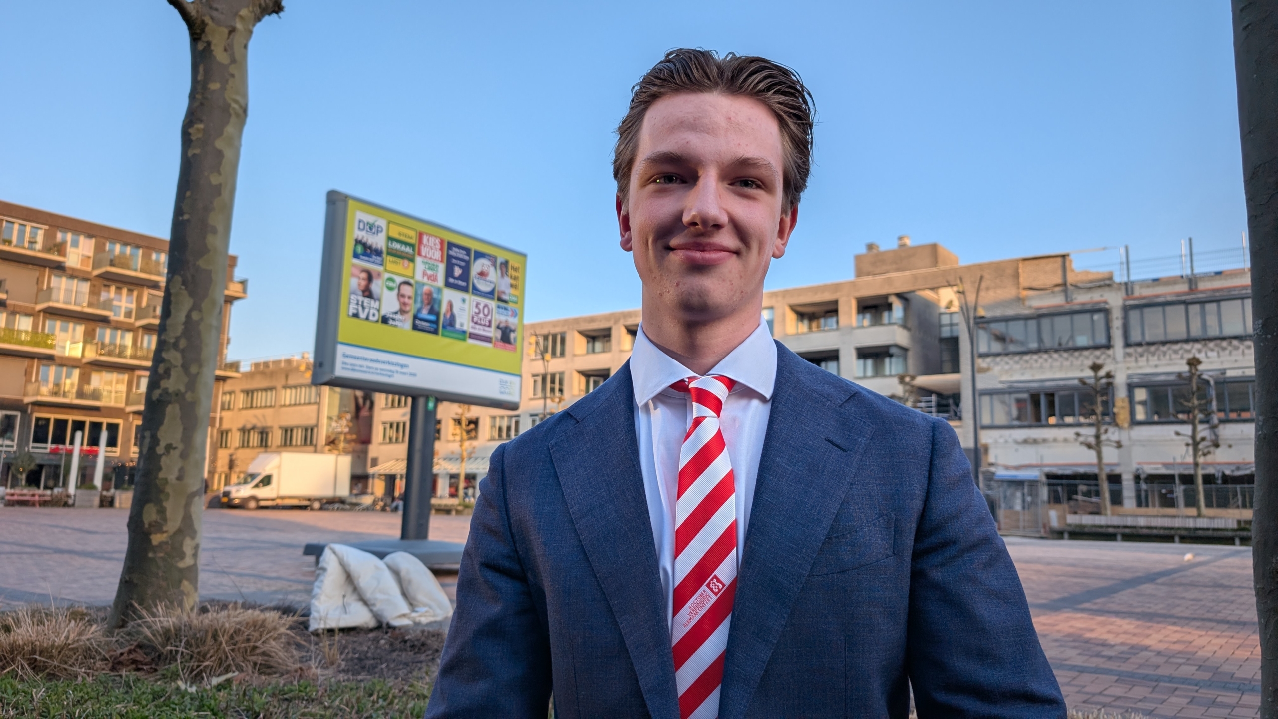 Jonge man in een pak met een gestreepte das staat in een stedelijke omgeving met verkiezingsborden op de achtergrond.