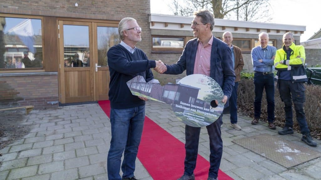 Twee mannen schudden handen en houden een grote symbolische sleutel vast, omringd door toeschouwers voor een gebouw.