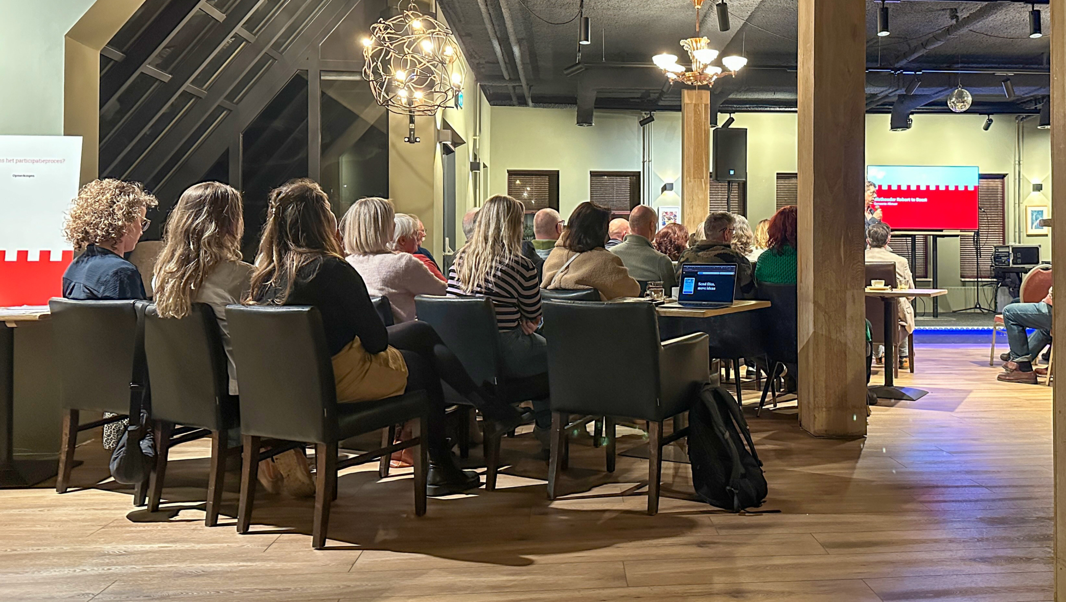 Een groep mensen luistert aandachtig naar een presentatie in een moderne zaal met houten elementen en gedempt licht.