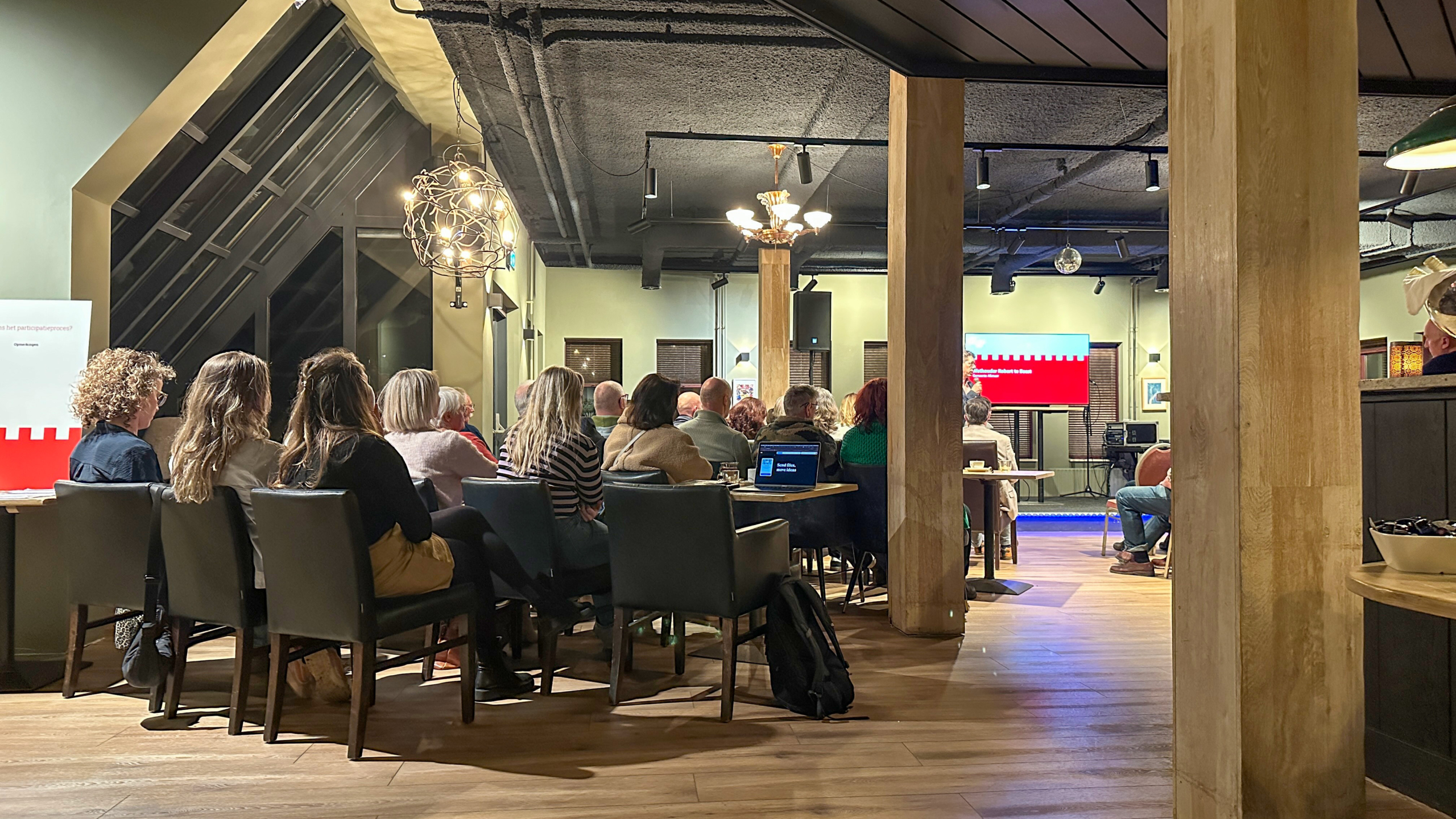 Een groep mensen luistert aandachtig naar een presentatie in een moderne zaal met houten elementen en gedempt licht.