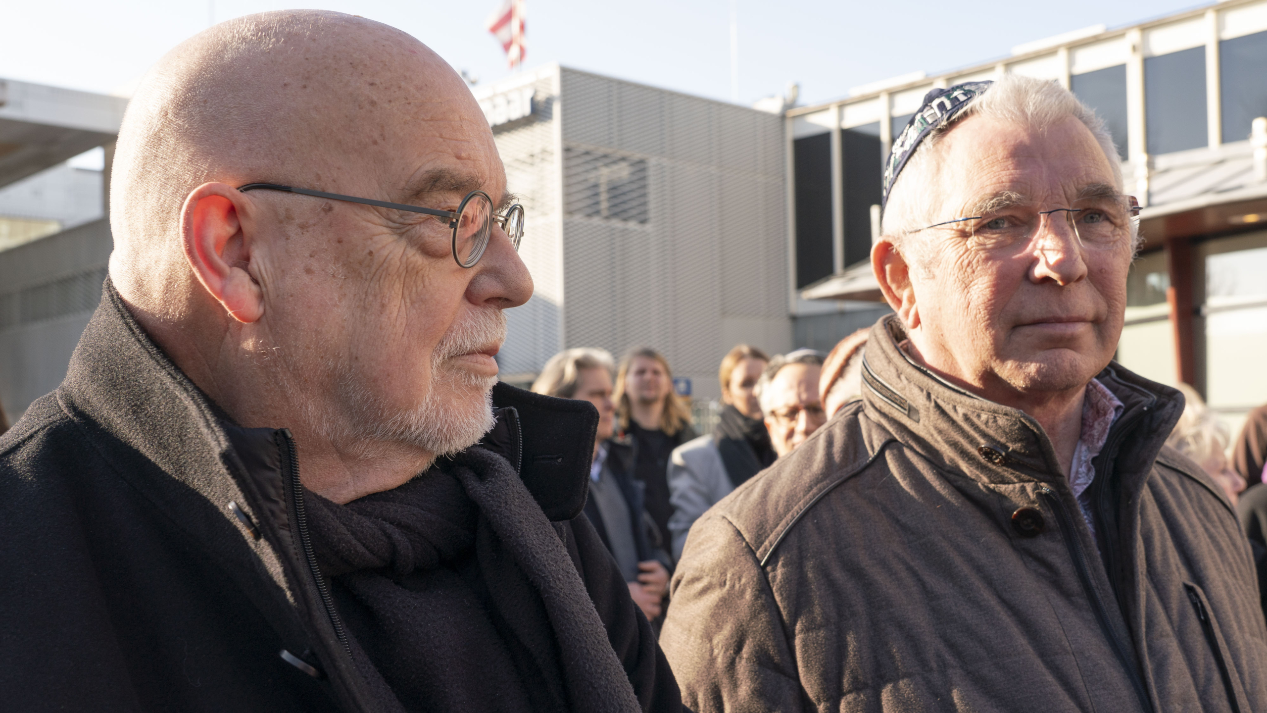 Twee oudere mannen staan buiten, beiden dragen brillen en warme jassen, omringd door een groep mensen.