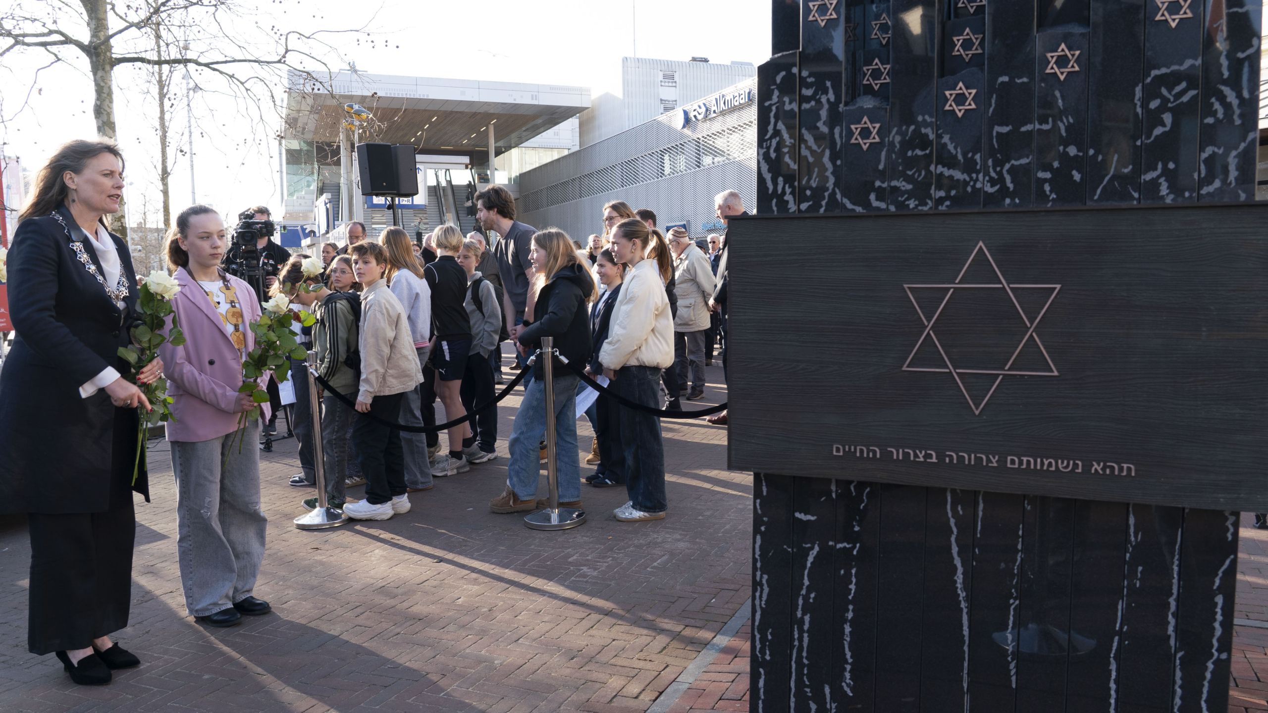 Groep mensen, waaronder een vrouw en een meisje met bloemen, voor een monument met davidsterren tijdens een herdenkingsdienst.