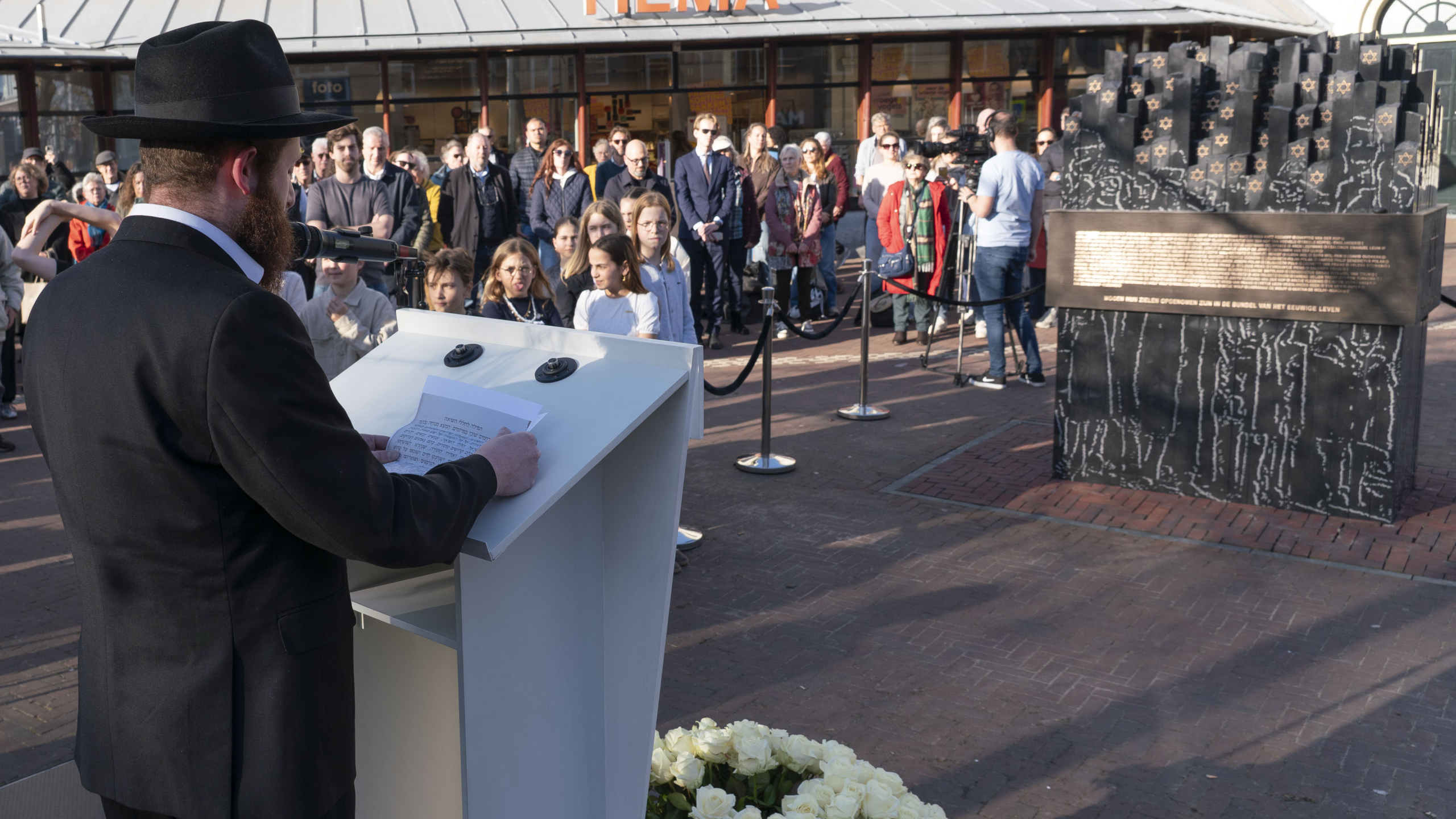 Een man in een zwart pak houdt een toespraak bij een herdenkingsmonument, terwijl een groep mensen toekijkt.