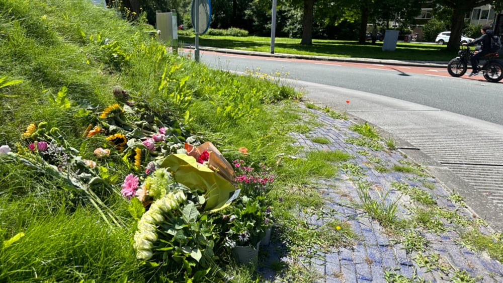 Bloemen leggend langs een weg, mogelijk een herdenkingsplek, met op de achtergrond een fietser op een zonnige dag.