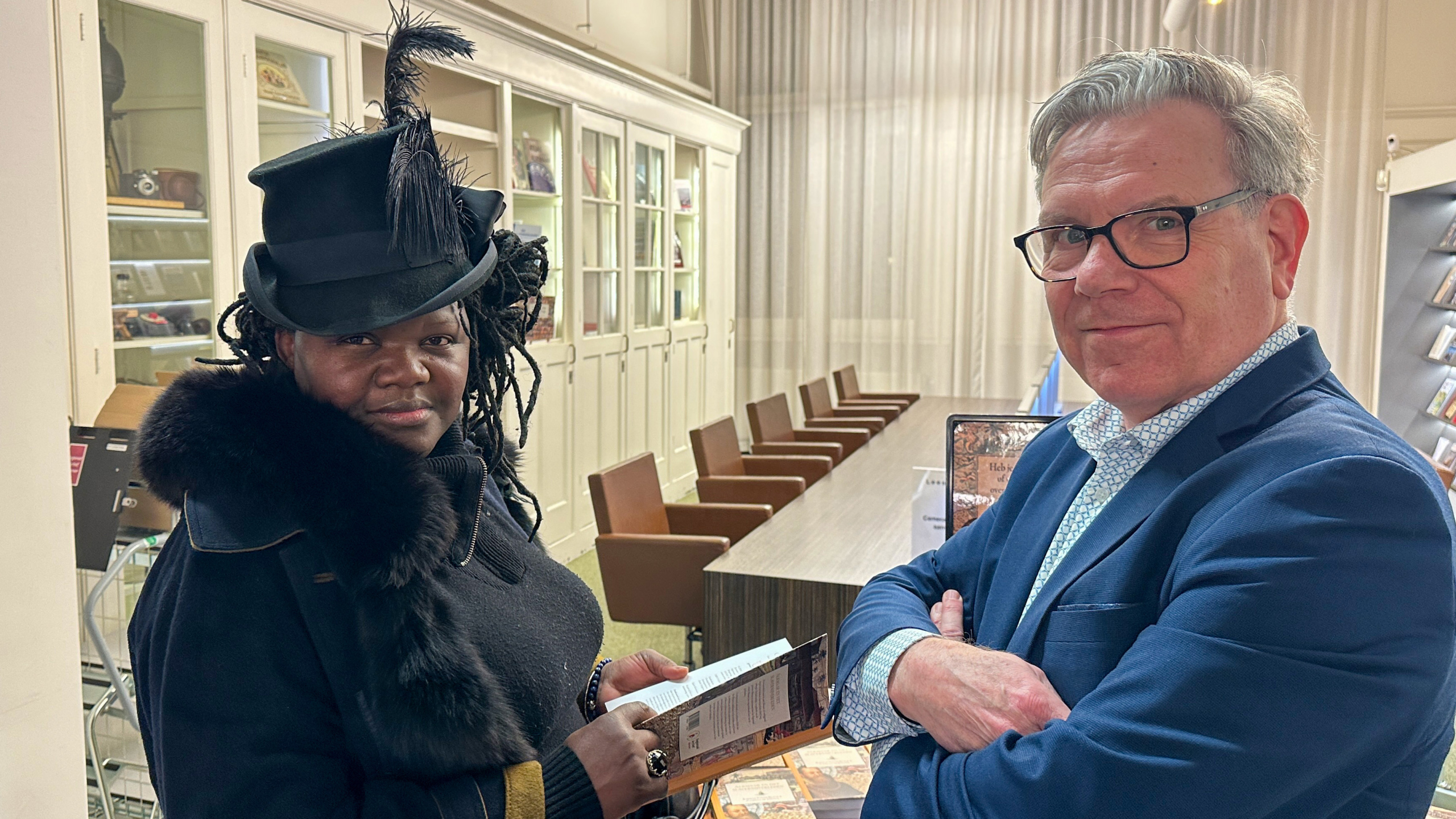 Twee mensen poseren in een kamer met een tafel en boekenplanken; een draagt een hoed met veren.