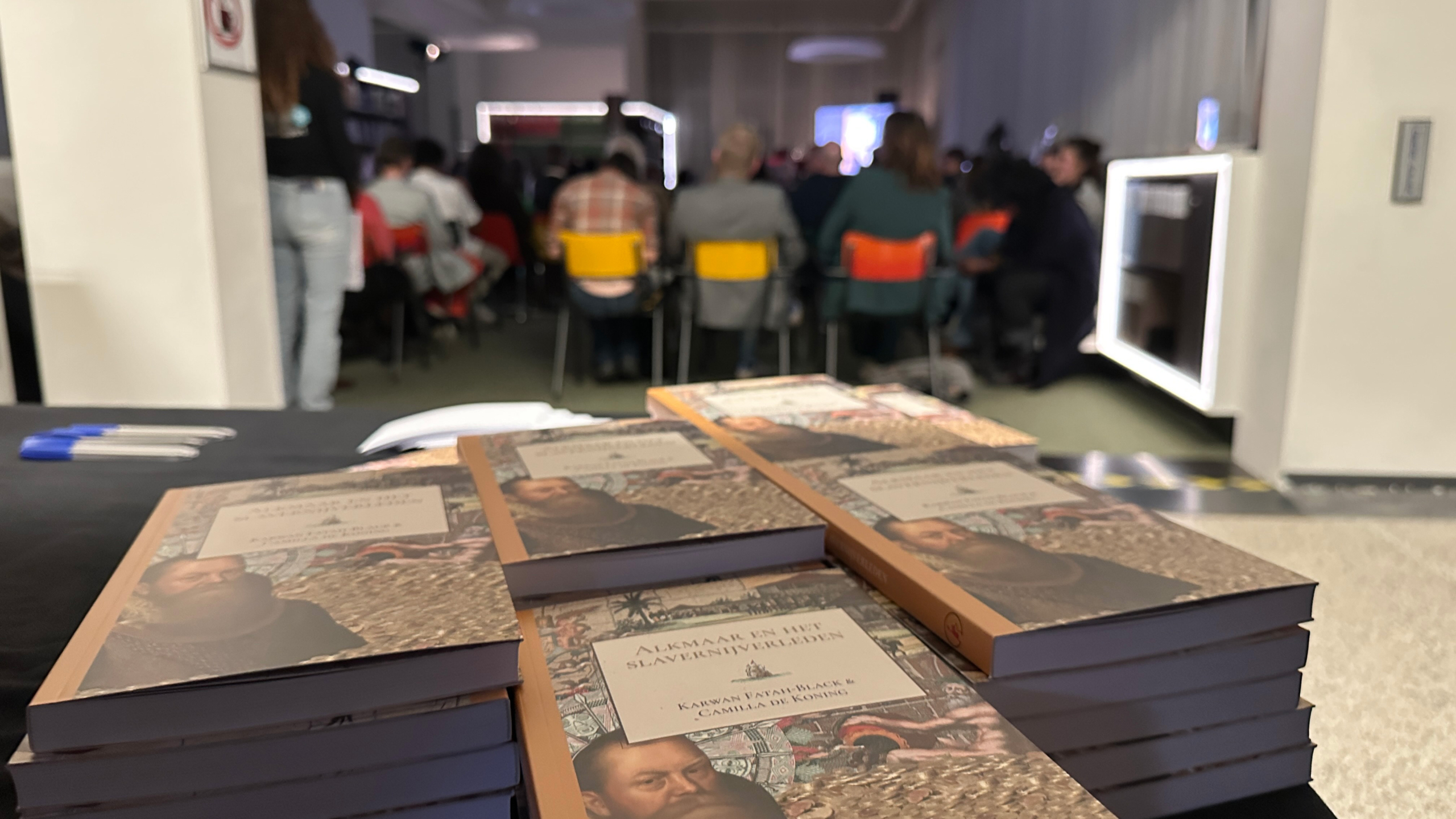 Stapels boeken op een tafel in een zaal met mensen zittend op kleurrijke stoelen, bij een evenement.