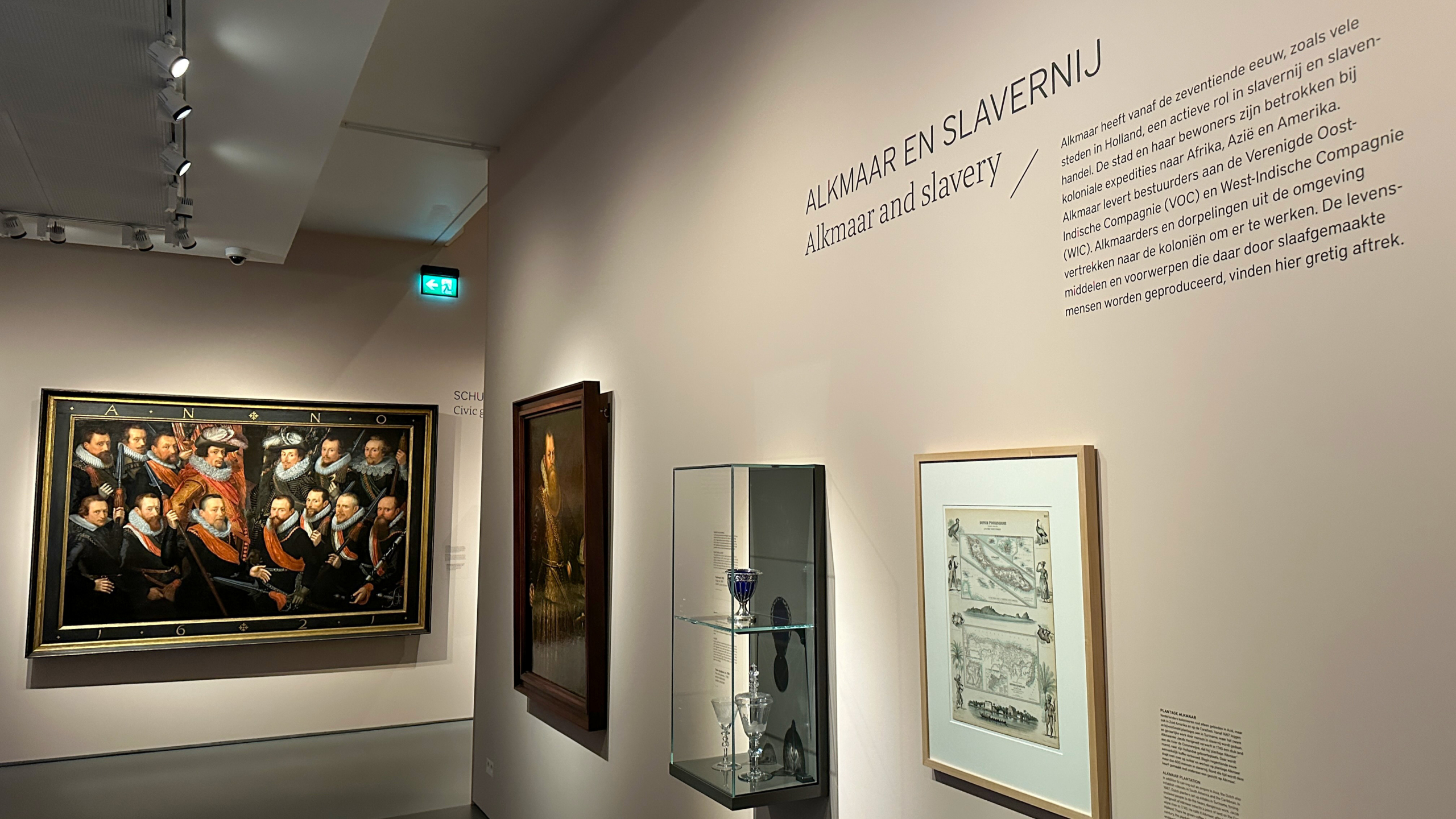 Museumexpositie over Alkmaar en slavernij met schilderijen en glazen vitrines, waaronder tekst op de muur met een uitleg.