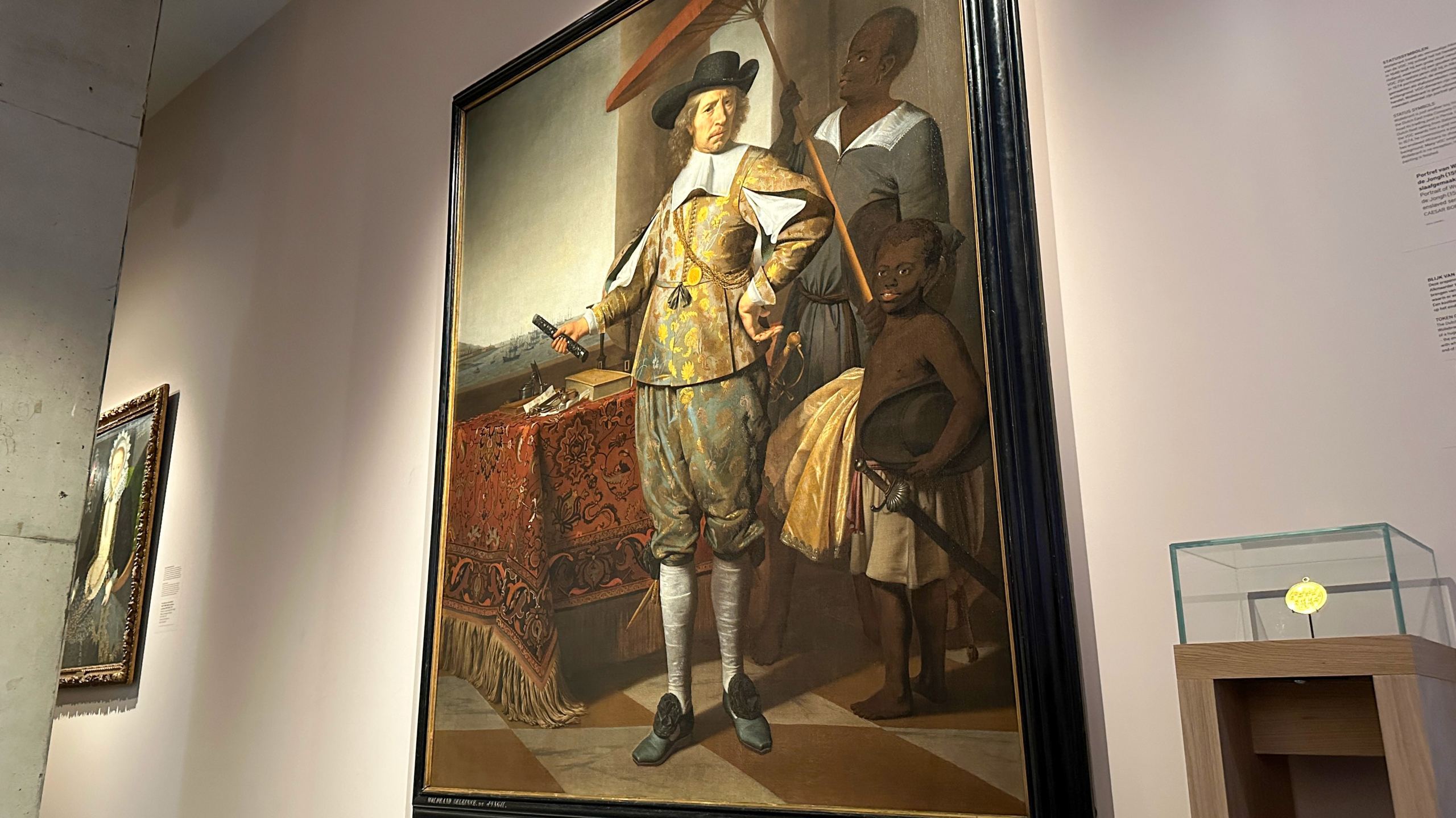 Schilderij in museum toont een man in 17e-eeuwse kleding met twee bedienden, in een interieur met een gedetailleerd tafelkleed.