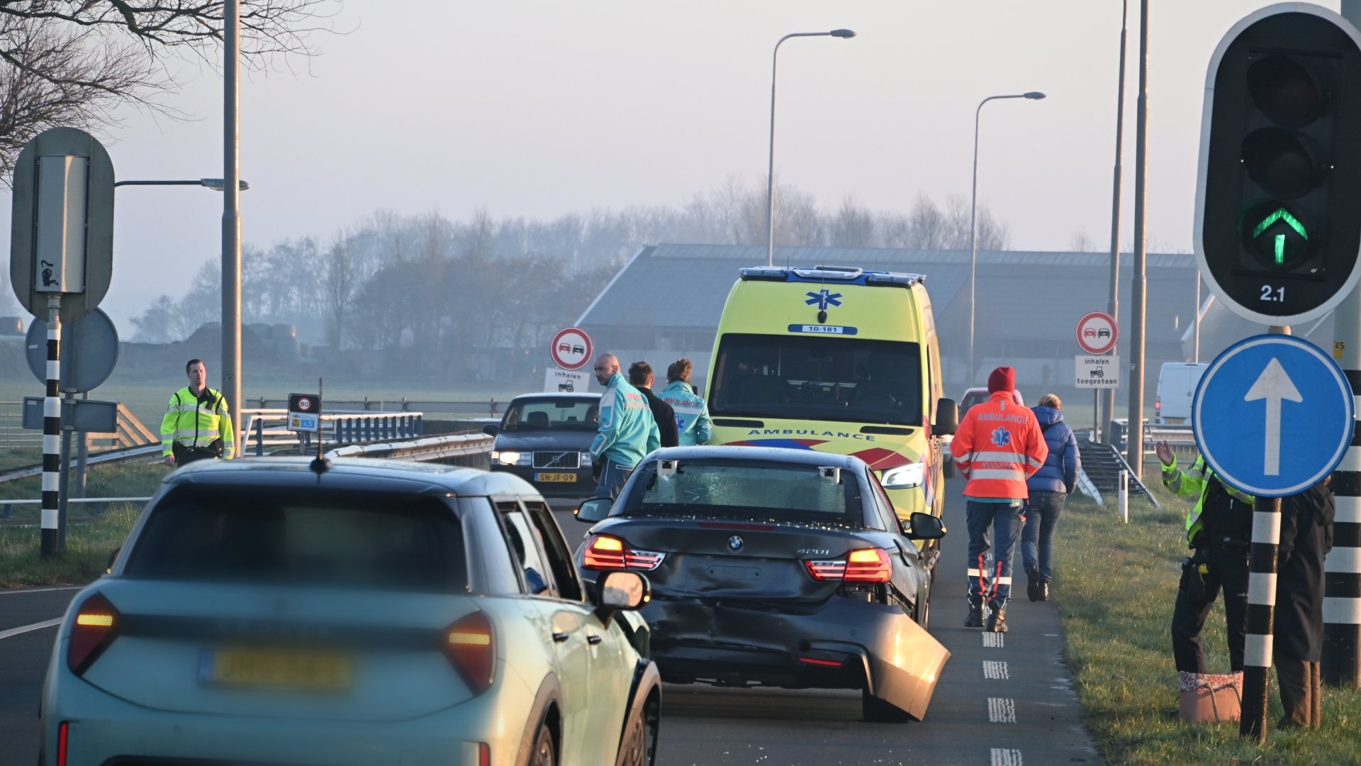 Auto-ongeluk met een ambulance en hulpverleners op de weg, waarbij meerdere auto's en verkeersborden zichtbaar zijn.