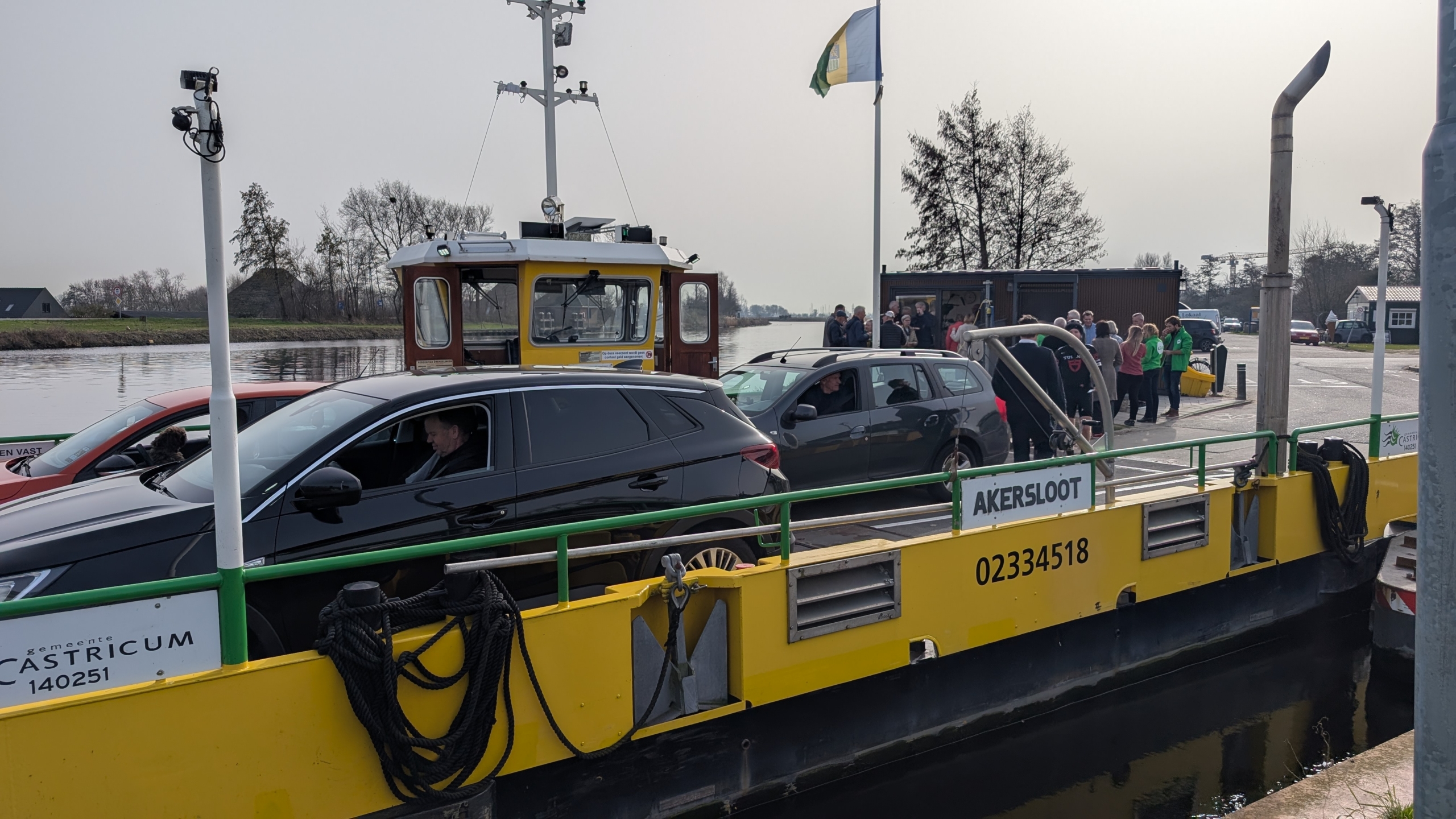 Auto's staan op een veerpont genaamd Akersloot, met op de achtergrond mensen en een rivier.