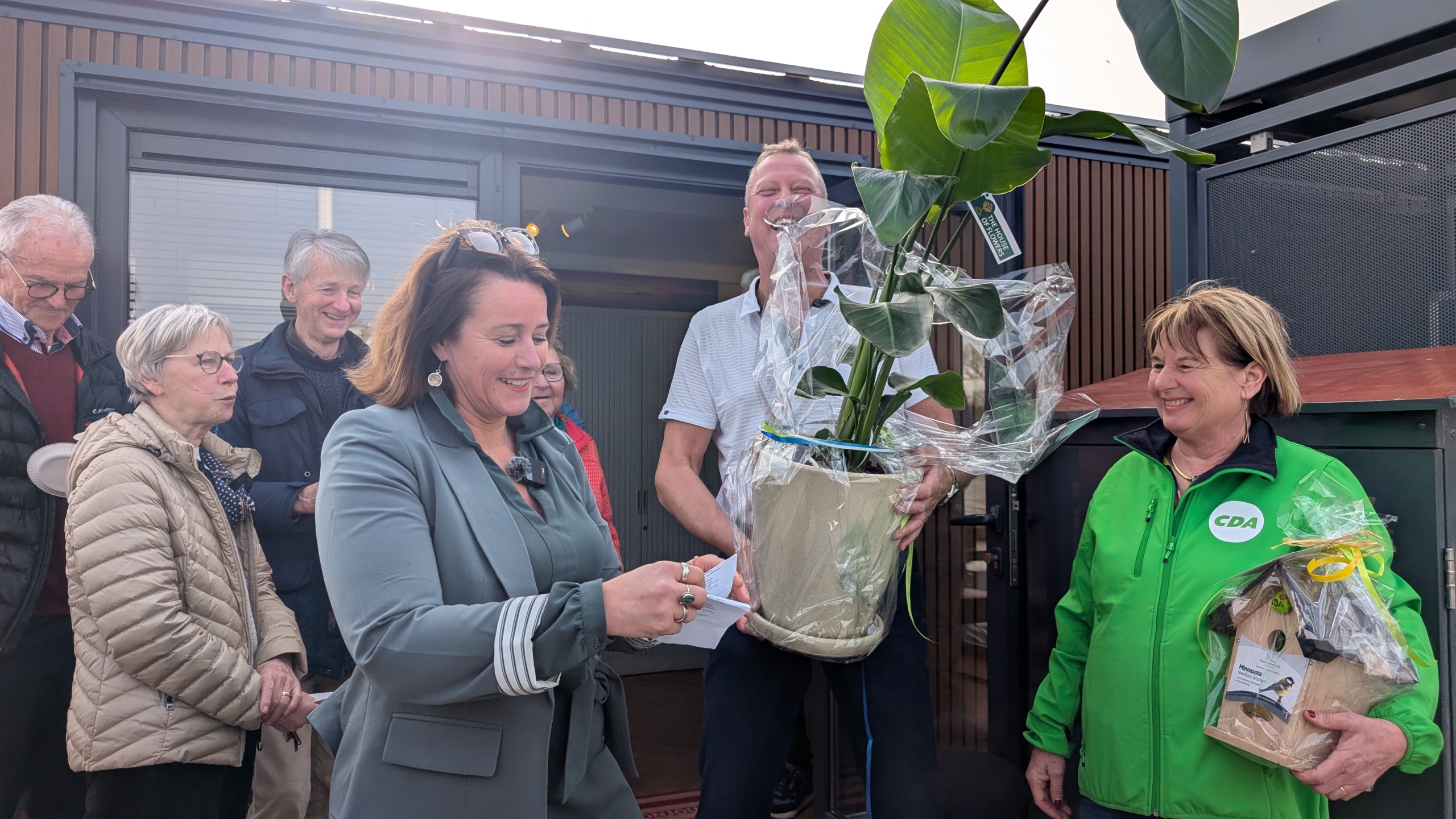 Groep mensen buiten, een vrouw opent een cadeau, en een andere persoon houdt een potplant vast. Een vrouw draagt een groene jas met CDA-logo.