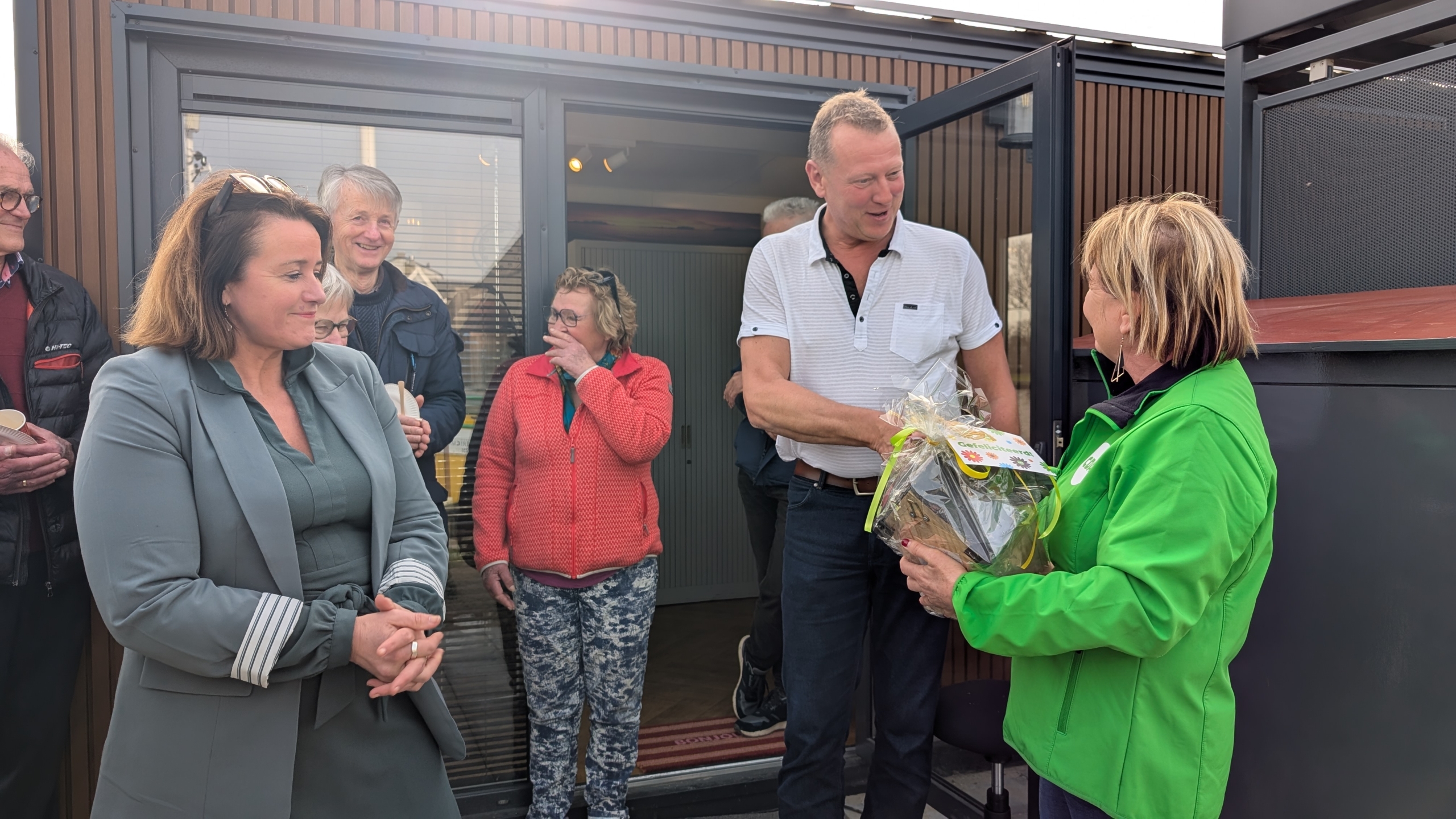 Een vrouw in een groene jas overhandigt een cadeau aan een man omringd door een groep mensen buiten een gebouw.